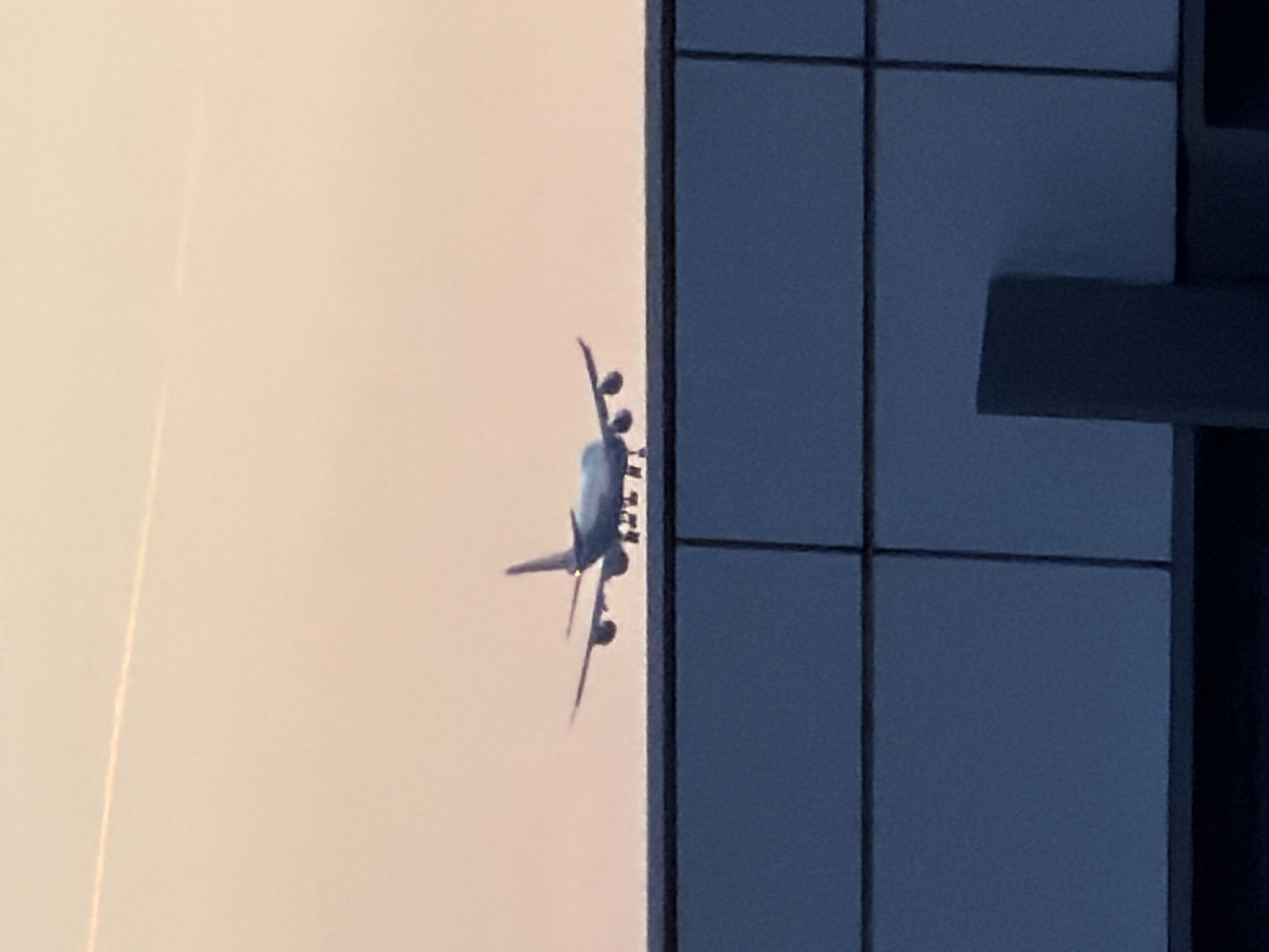 A passenger airplane is captured in mid-air, appearing to be in the process of landing or taking off, with its landing gear extended. The aircraft is silhouetted against a soft, pale orange sky, suggesting either sunrise or sunset. A faint contrail is visible in the upper portion of the sky. In the foreground, a dark, textured structure, likely a building or barrier, is partially visible. The image appears to have been taken from a low vantage point, looking upwards towards the aircraft. The setting is within the municipality of Leudelange, Luxembourg.