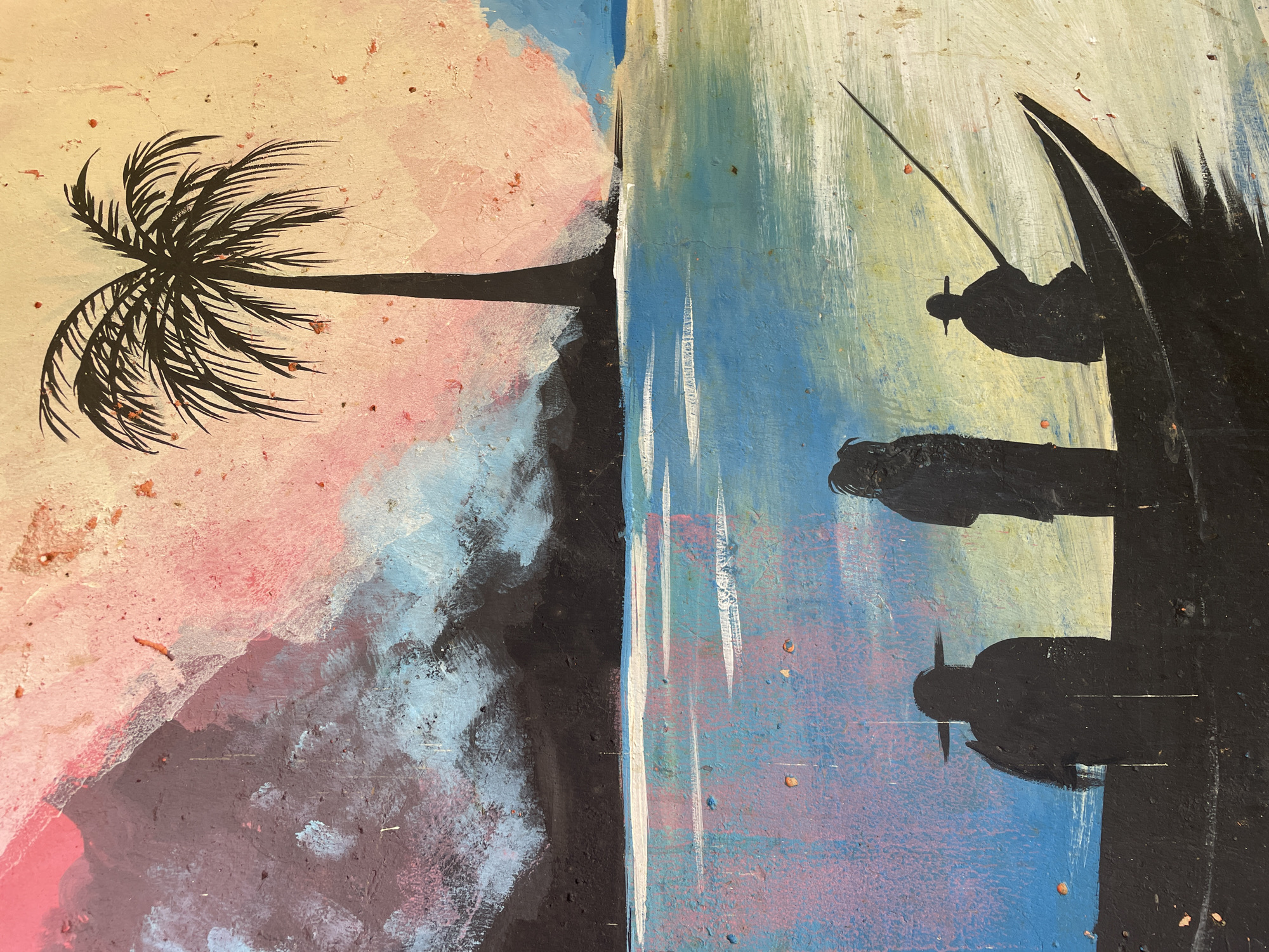 This media file is a painting or mural depicting a stylized tropical or coastal scene, likely rendered on a textured surface such as a wall or rough canvas. The painting utilizes bold silhouettes against an impressionistic background.
On the left side of the composition, a prominent black silhouette of a palm tree stands against a vibrant sky. The tree features a long, slender trunk extending towards the center, with its numerous fronds detailed and appearing to be swayed by a breeze. The sky behind the palm tree is painted in soft, blended hues of pale yellow, pink, and light blue, suggesting either dawn or dusk.
A distinct, thick black line or barrier separates the land with the palm tree from the body of water and figures on the right. The water is depicted with broad, dynamic brushstrokes in shades of blue, teal, and hints of purple and pink, giving it a luminous, reflective quality. Vertical white strokes interspersed in the water suggest movement or light reflections.
On the far right, three black human silhouettes are positioned on what appears to be a dark landmass, possibly a shoreline or a boat. The uppermost figure is actively engaged in fishing, holding a long, thin fishing rod extending into the water. This figure wears a distinct, possibly conical, hat. Below this, two other, less detailed, human silhouettes are visible, also appearing to be seated or standing and observing the water.
The overall mood is tranquil and contemplative, with the silhouetted forms creating a strong contrast against the colorful, atmospheric background. The textured surface of the painting is noticeable throughout, with small reddish-brown flecks and irregularities adding to its rustic charm. There is no visible text in the image.
