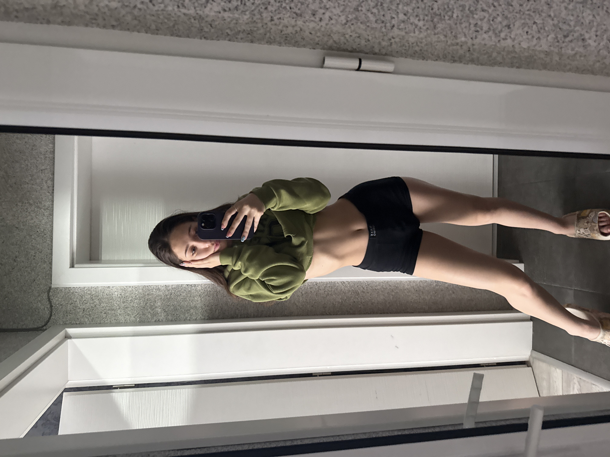 This image is a full-body mirror selfie of a young woman, captured indoors and rotated 90 degrees counter-clockwise. The setting is an interior space, likely in Sofia, Bulgaria, characterized by modern design elements.
The woman has long, dark hair and is wearing a green, cropped hooded sweatshirt that reveals her midriff, paired with black shorts. On the shorts, mirrored text that appears to be "XXAM OMOO" is visible. She is wearing light-colored, patterned slip-on sandals. Her left hand holds a dark-cased smartphone (possibly purple) to take the picture, partially obscuring her face, while her right hand rests near her cheek. Her fingernails are painted a light blue.
The environment reflected in the mirror includes a light grey, speckled carpeted floor transitioning to a darker grey tiled floor. White door frames or window frames are also reflected, outlining the boundaries of the room. The lighting appears bright and even, suggesting either natural daylight or well-distributed artificial light. The woman's pose is casual and relaxed as she captures her reflection.