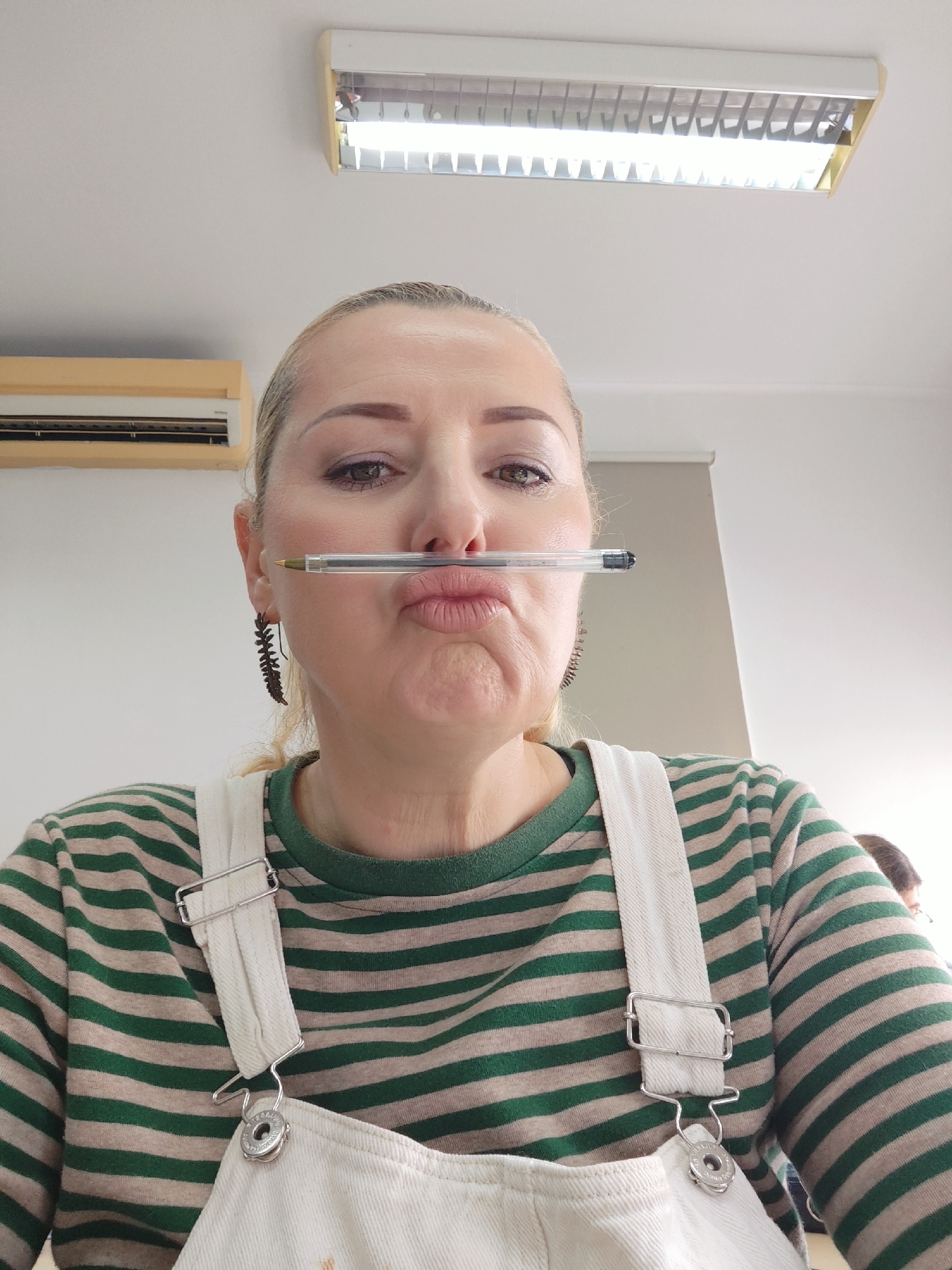 A medium close-up, eye-level shot captures a woman indoors, facing the camera with a playful expression. She is balancing a clear ballpoint pen horizontally between her upper lip and nose, her lips pursed in a humorous way. The woman has light blonde hair, pulled back from her face, and light-colored eyes. She wears makeup, including visible eyeliner. A dark, decorative earring, shaped like a fern or leaf, dangles from her left ear (viewer's right).
She is dressed in a long-sleeved shirt with alternating green and light beige horizontal stripes. Over this, she wears light-colored (likely white or off-white) overalls, with silver-toned metal buckles and fasteners visible on the shoulder straps.
The background consists of plain, light-colored walls and a white ceiling, typical of an indoor environment. On the ceiling, a long, rectangular fluorescent light fixture emits a bright glow. To the upper left, another lighter-colored rectangular unit, possibly an air conditioner or heater, is mounted on the wall. No discernible text is visible on any objects or surfaces. The overall scene conveys a lighthearted and humorous moment, captured indoors, potentially in Ponta Delgada, Portugal.