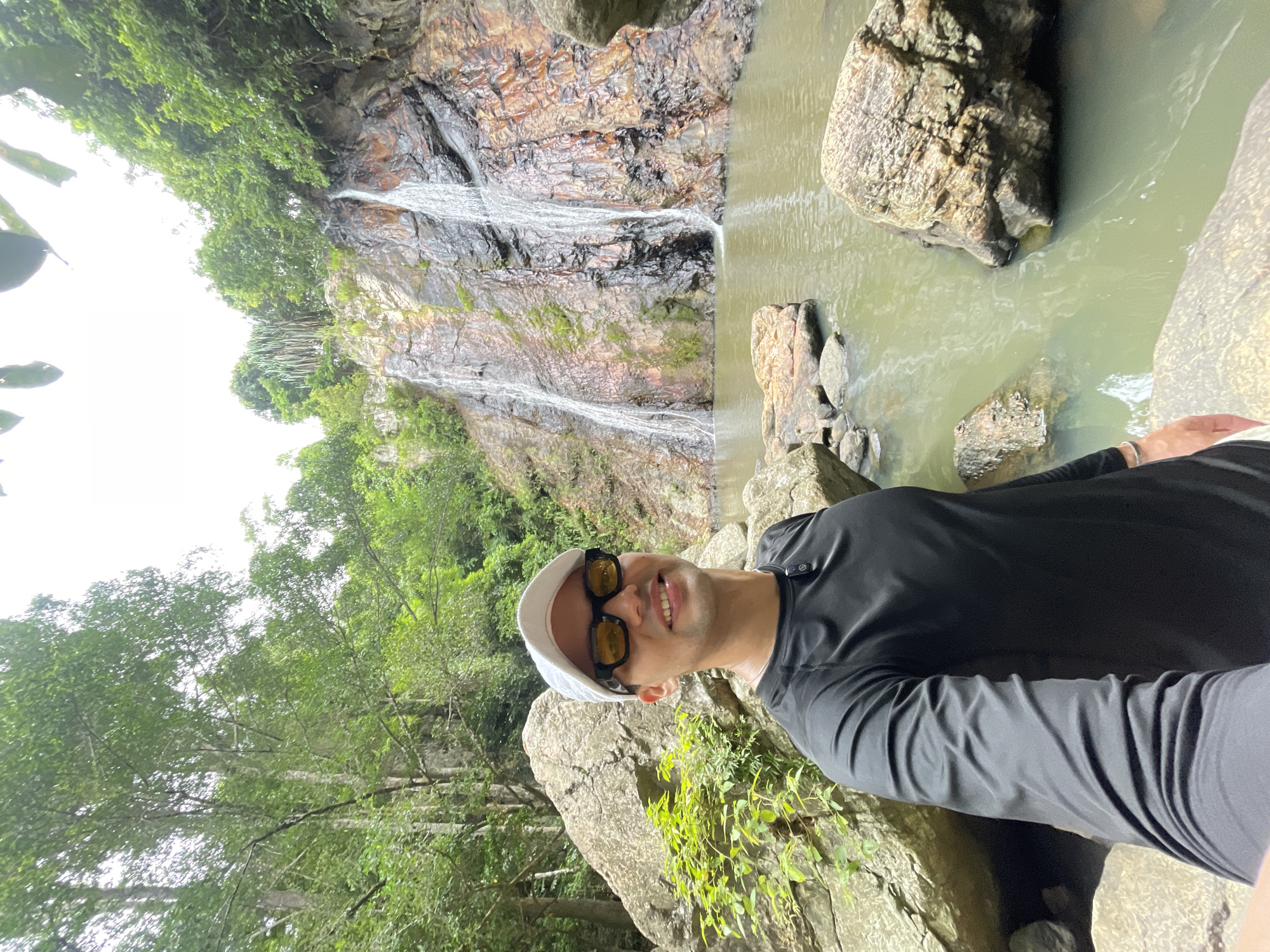 A young man in a black long-sleeved rash guard, white baseball cap, and yellow-tinted sunglasses is taking a selfie in front of a two-tiered waterfall in Baan Thurian, Thailand. The waterfall cascades down a rocky cliff face covered in moss and vegetation. The water collects in a calm, greenish pool at the base. Large, weathered boulders are scattered around the pool and the surrounding area, which is densely forested with lush green trees and foliage. The man is smiling and appears to be enjoying the natural scenery.