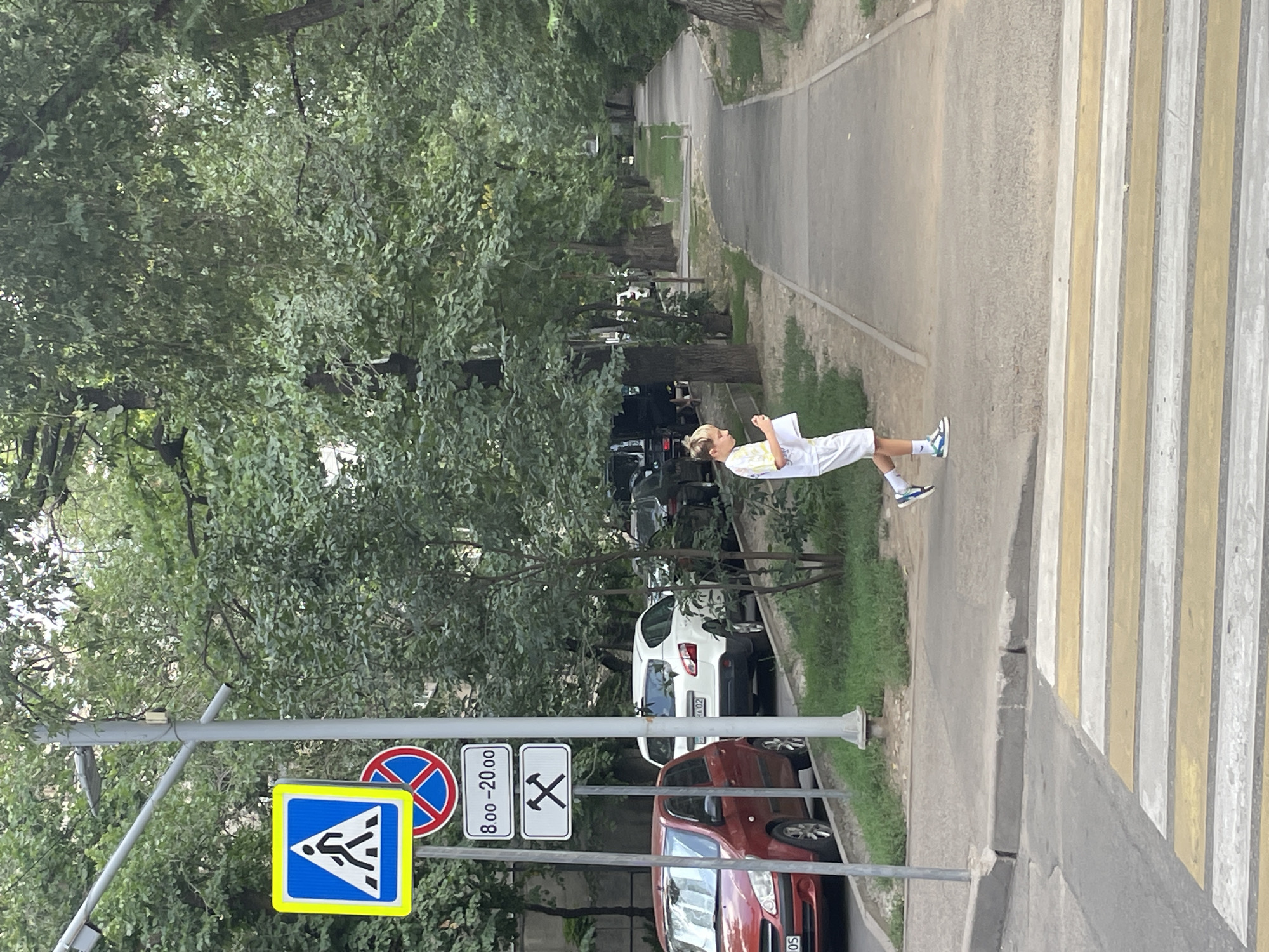 The image depicts a street scene in Almaty, Kazakhstan, featuring a pedestrian crossing and a sidewalk lined with trees. In the foreground, a boy in white shorts and a t-shirt is walking on the sidewalk, holding a piece of paper. He is wearing athletic shoes. A red car is parked on the left side of the street, and other vehicles are visible further down the road. A utility pole with several traffic signs is also present, including a pedestrian crossing sign, a prohibitory sign, and signs indicating working hours and possibly a no-parking zone. The environment is characterized by mature trees providing shade, suggesting a warm day. The sidewalk is paved, and the street has a zebra crossing painted on it. The overall scene is a typical urban environment with pedestrian infrastructure and vehicular traffic.