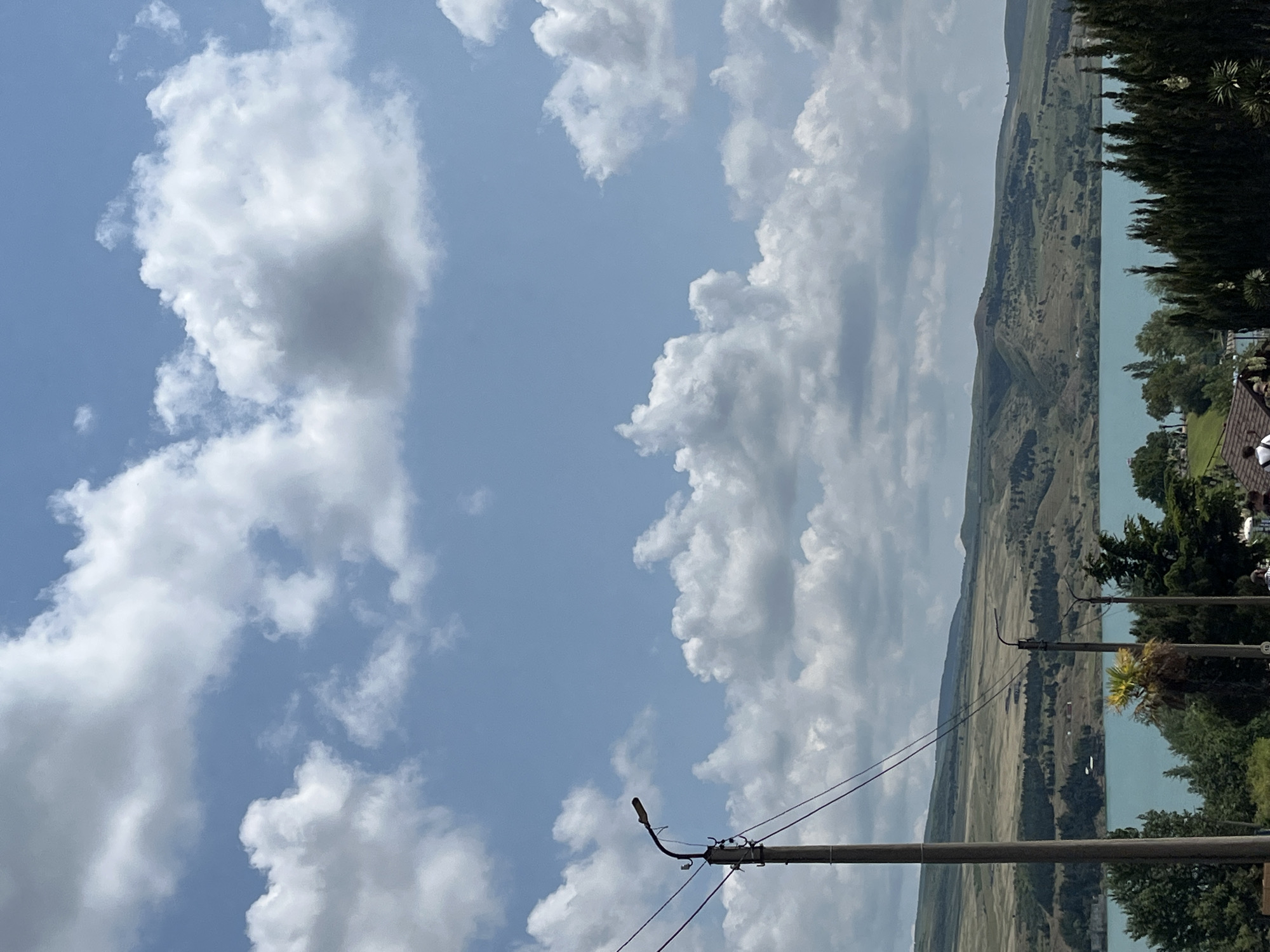 The image depicts a scenic landscape with a body of water in the middle ground, flanked by rolling hills and a clear blue sky filled with fluffy white clouds. In the foreground, a tall wooden utility pole with power lines stretches upwards. The water appears to be a reservoir or lake, with the undulating hills behind it covered in greenery and scattered structures. The sky is bright, suggesting daylight, and the overall scene conveys a sense of natural beauty and tranquility. There are no people or specific city landmarks identifiable in the image that would allow for pinpointing a precise location within Tbilisi, Georgia.