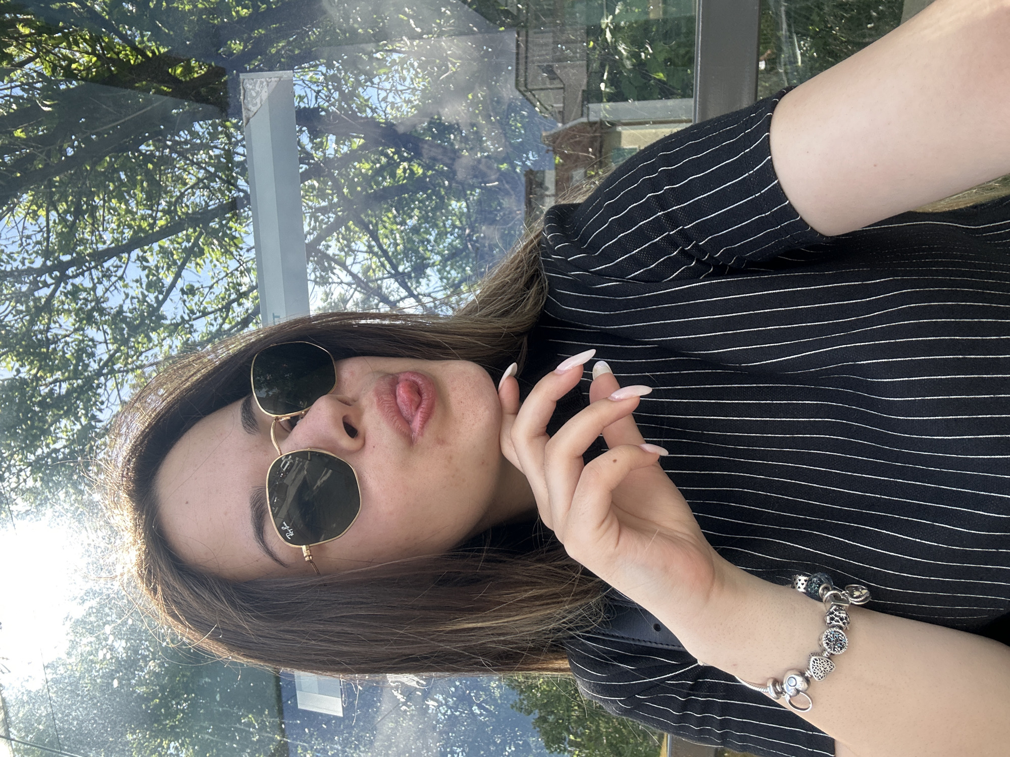 A woman with long brown hair is posing for a photo outdoors. She is wearing sunglasses and a black and white striped shirt. She is holding her hand up to her face and making a kissing face. Her right arm is visible and she has a silver Pandora bracelet on her wrist. Behind her is a glass panel, reflecting trees and sky, as well as what appears to be a building structure. The exact location in Sofia, Bulgaria cannot be determined from the image alone.