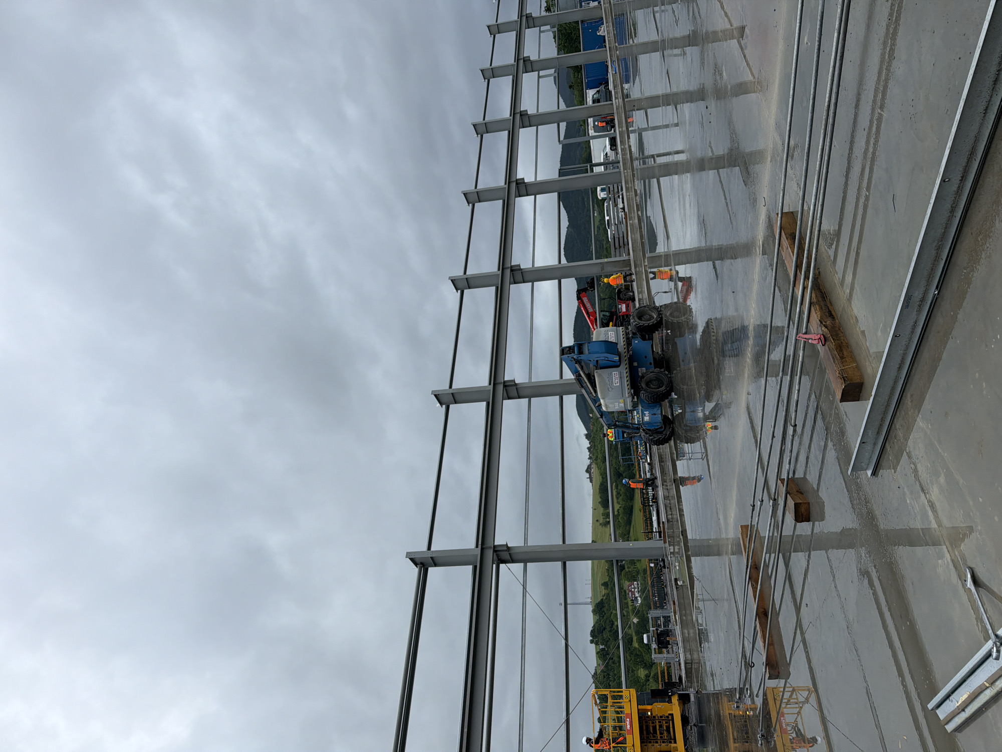 This is an exterior, eye-level, medium shot of a construction site in Janowice, Poland. The scene is dominated by the skeletal framework of a large building under construction, composed of grey steel beams supported by vertical columns. The ground is a wet, reflective concrete surface, indicating recent rain or damp conditions. Several construction workers, wearing high-visibility vests and hard hats, are present. Two workers are operating mobile elevated work platforms, one yellow and one blue, which are positioned near the steel structure. A red crane is also visible in the background. In the distance, rolling green hills and trees frame the construction site under a cloudy, overcast sky. The overall atmosphere is one of ongoing construction work in a somewhat rural, hilly environment.