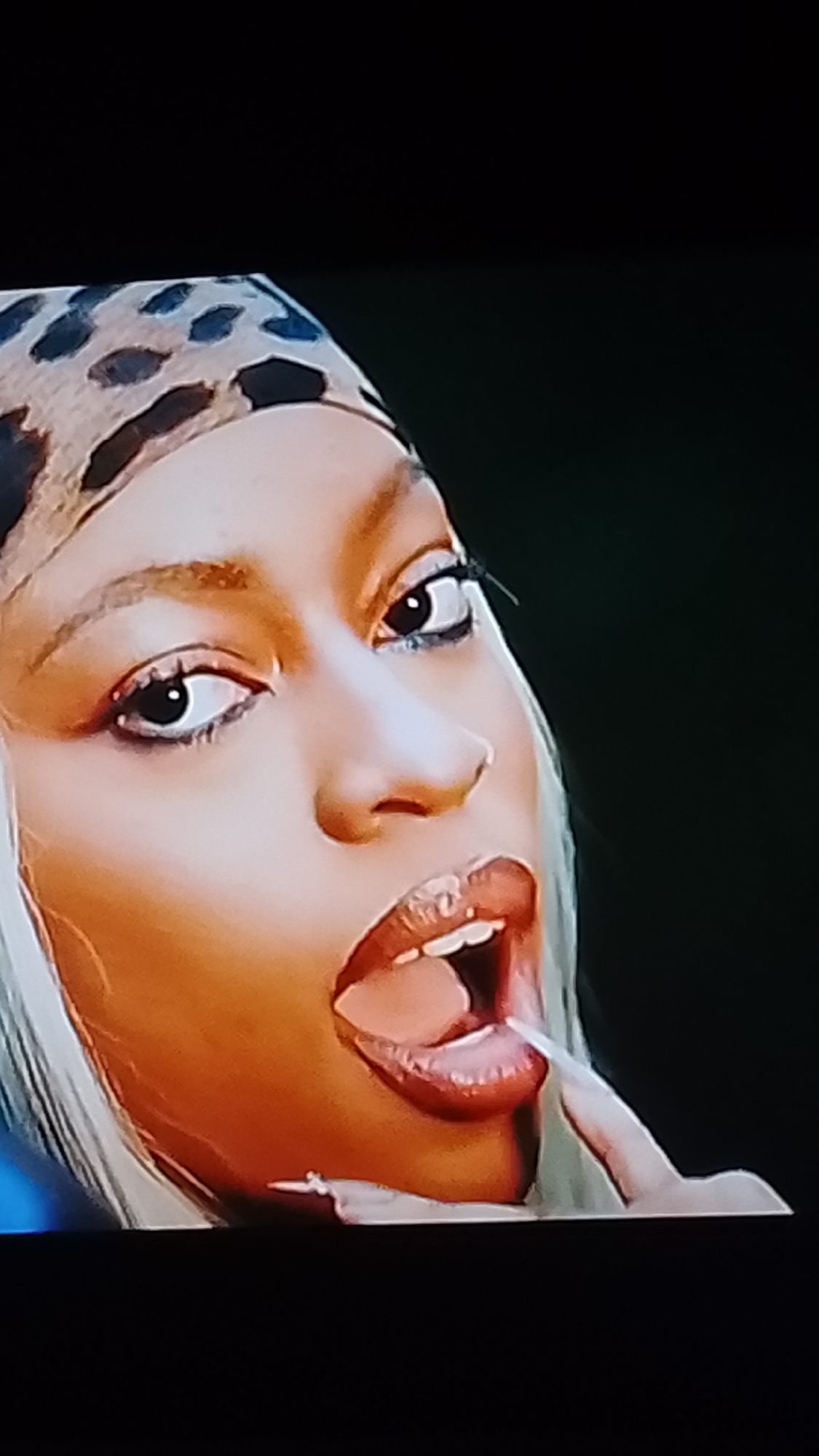 The image is a close-up shot of a woman's face, angled to the right. She has light skin and is wearing a leopard print headscarf that covers the top of her head. Her eyes are open and looking to the left. Her mouth is open, with her tongue visible. She appears to be touching her tongue with her finger, which has long, light-colored fingernails. Her lips are full and glossy, with dark lipstick. Her eyebrows are defined and arched. Her hair is long and blonde, peeking out from under the headscarf. The background is dark and out of focus. There is no discernible indication of the location being in Lokoja, Nigeria, or any other specific city. The scene appears to be a posed shot, possibly for a photoshoot or a music video.