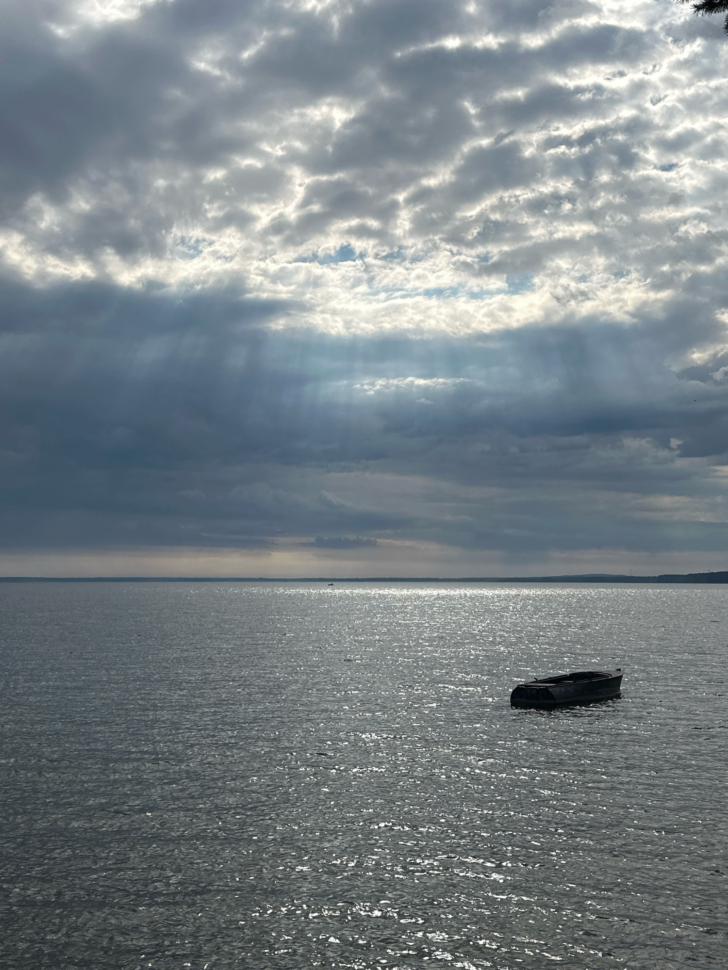 The image depicts a wide expanse of calm water, likely a lake or large river, under a dramatic, cloudy sky. Rays of sunlight pierce through gaps in the clouds, creating a dappled effect on the water's surface. In the foreground, a solitary, dark-colored rowboat floats gently. The shoreline is visible in the distance as a faint, dark line against the horizon, with a hint of greenery suggesting land. The overall atmosphere is serene yet moody, with the play of light and shadow on the water and in the sky being a prominent feature. The location is identified as Нарацкі сельскі Савет, Belarus.