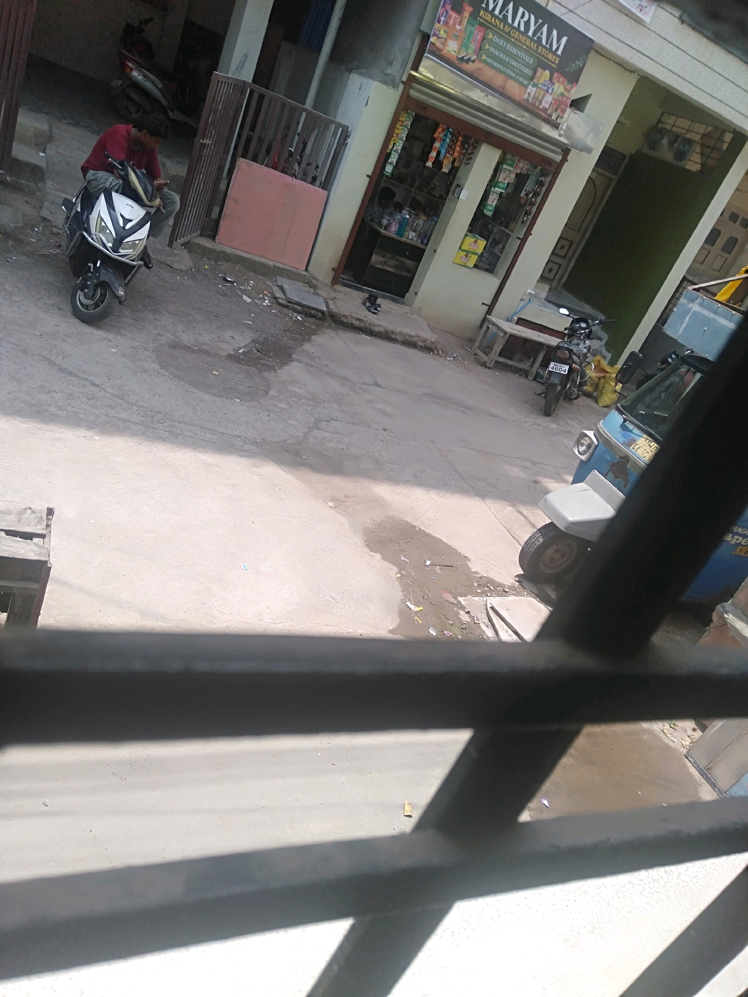 A man in a red shirt sits on a white and black electric scooter in front of a store. The scooter is parked on a paved street. The store is named "MARYAM" and appears to be a kirana and general store. A blue auto-rickshaw is parked to the right of the store, and a motorcycle is parked in front of the store. The scene is likely in Shirpur, India, based on the provided context. The street is somewhat dirty, with some puddles of water and debris. The image is taken from an elevated perspective, looking down at the street and the scene.