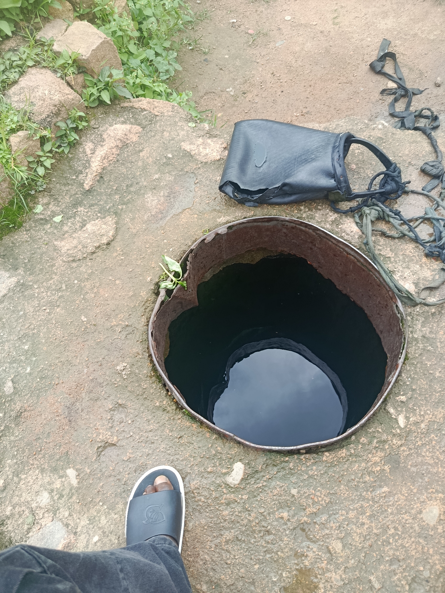 An overhead, medium shot shows a person's foot wearing a black sandal and dark pants on a damp, rocky surface. To the right of the foot, a rusty metal rim surrounds a dark opening filled with water, resembling a well or a cistern. Adjacent to the opening, a black leather bag with frayed straps and a rope lies on the ground. In the upper left corner, large rocks are scattered among green vegetation and moss. The overall scene suggests an outdoor, possibly rural or undeveloped, location.