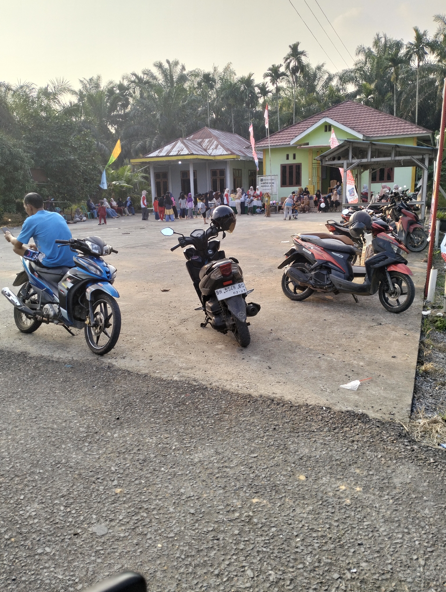 In Mandala Jaya, Indonesia, a street scene unfolds with a focus on a group of people gathered outside a light green building with a red-tiled roof, identified by signage as "KANTOR DESA MANDALA JAYA". The building appears to be a community center or government office. Several parked motorcycles are visible on a concrete paved area in front of the building, including a blue motorcycle in the foreground to the left, occupied by a person in a blue shirt facing away from the camera, holding a mobile phone. To the right of the blue motorcycle, a black motorcycle is parked. Further back, other motorcycles are parked, including a red and black scooter.
A crowd of individuals, including adults and children, are congregated in front of the building, some sitting on the concrete and others standing. Some are dressed in casual clothing, while others wear headscarves, suggesting a mix of genders and ages. Several Indonesian flags are displayed, including a large red and white flag on a pole to the left of the building. The environment is characterized by lush green foliage, including palm trees, indicating a tropical setting. The ground is a mix of asphalt and concrete. The lighting suggests it is late afternoon or early evening, with the sun casting a warm glow on the scene.