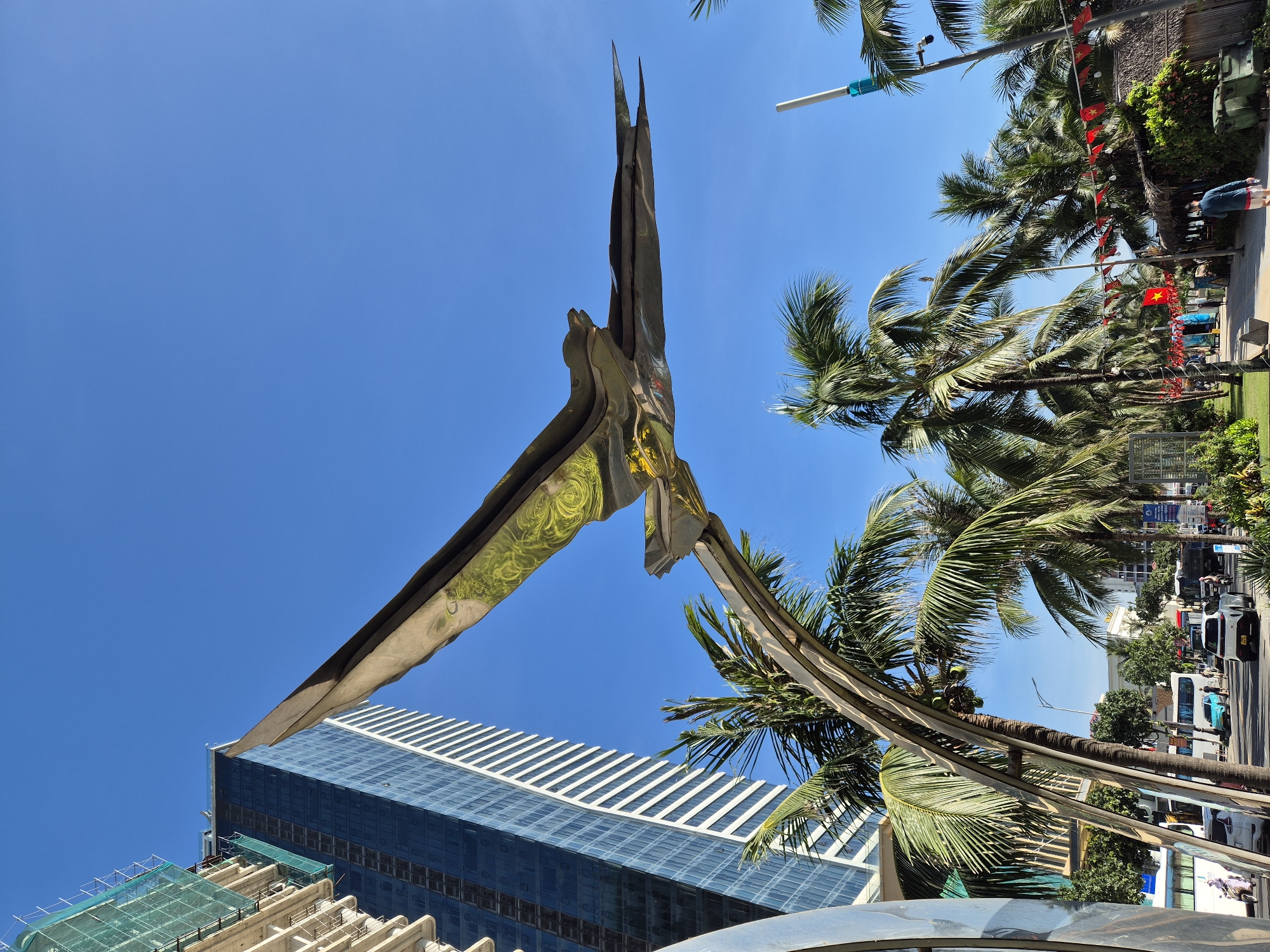 The image depicts a contemporary sculpture of a bird in flight, made of polished metal with reflective surfaces, positioned against a clear blue sky. The sculpture is situated in an urban environment, with a tall, modern glass-fronted skyscraper under construction in the background. Palm trees are abundant throughout the scene, suggesting a tropical or subtropical climate. In the foreground and midground, there are elements of a city street, including vehicles, traffic lights, and pedestrians. The presence of Vietnamese flags strung along a walkway indicates the location is likely Đà Nẵng, Vietnam. The overall impression is one of a vibrant, developing coastal city with a blend of modern architecture and natural tropical elements.