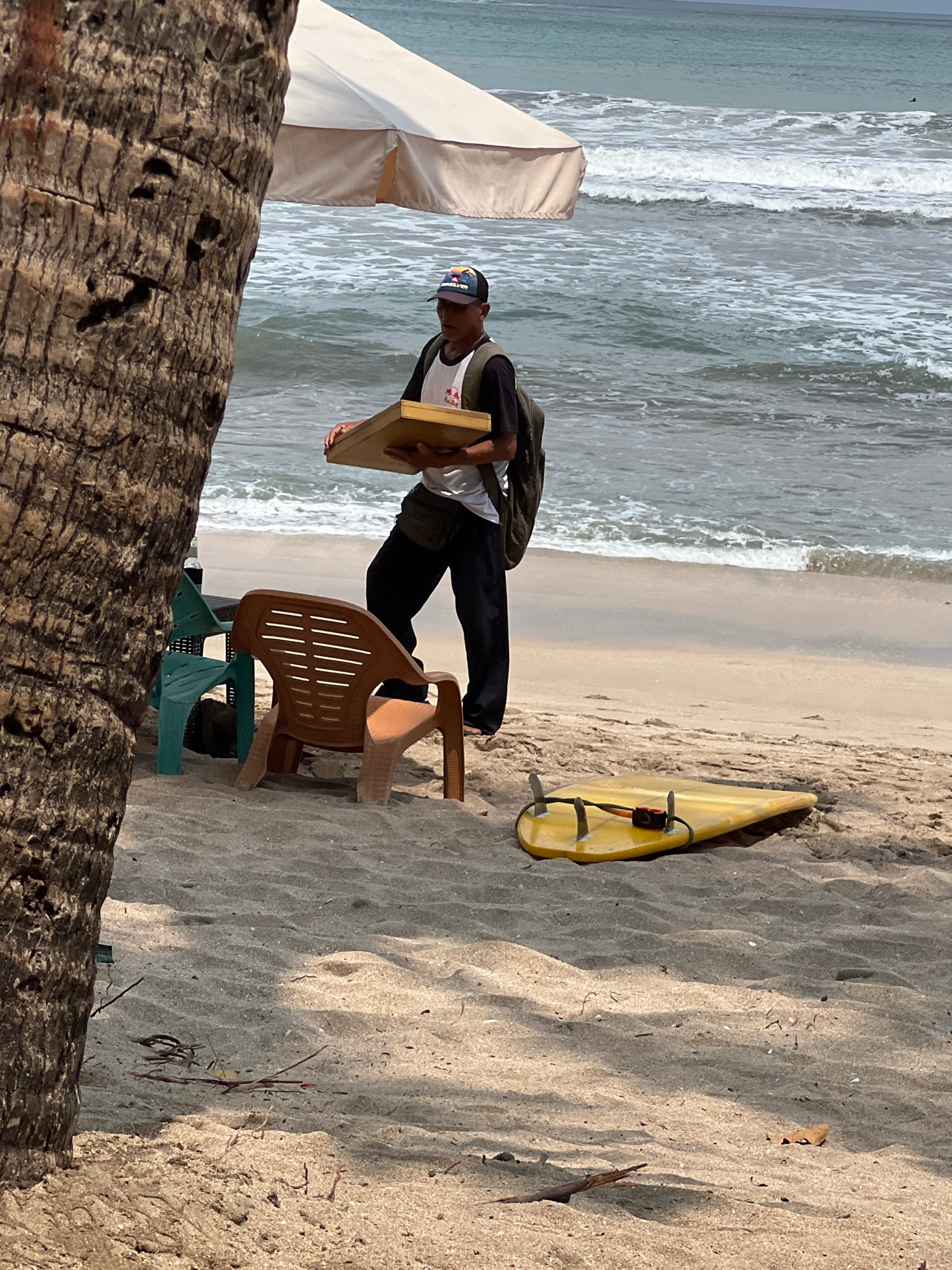 A man stands on a sandy beach in Kuta, Indonesia, holding a flat, rectangular object. He wears a dark baseball cap, a white t-shirt with a graphic design, black pants, and a backpack. In front of him, a yellow surfboard lies on the sand. Two chairs are positioned nearby, one a light brown plastic chair and the other a dark green plastic chair. A palm tree trunk is visible in the foreground on the left. Behind the man, waves crash on the shore under an overcast sky, and a light-colored beach umbrella is partially visible.