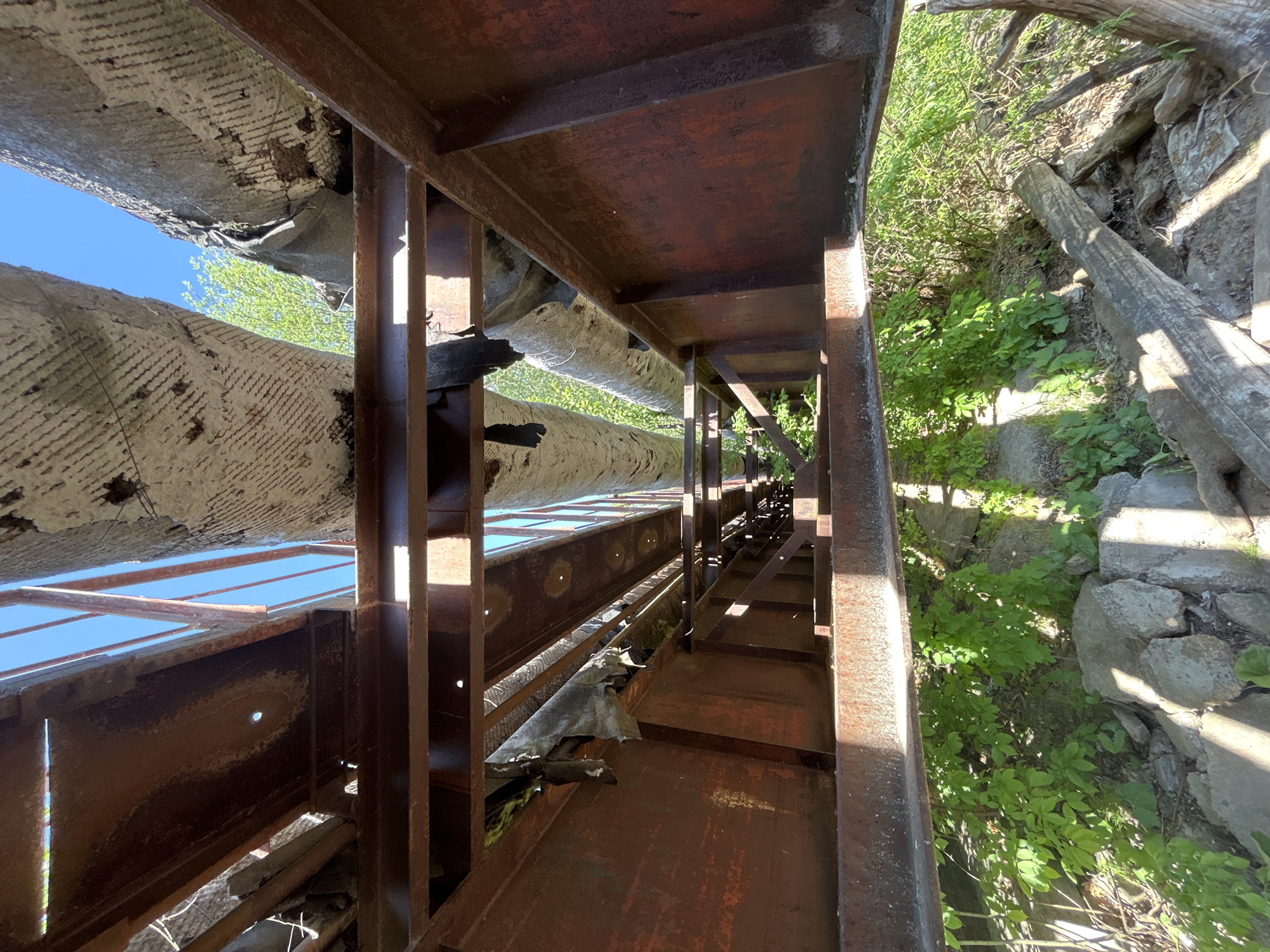 The image shows a rusted metal structure, likely a bridge or walkway, in what appears to be a state of disrepair. The structure is constructed of steel beams and platforms. Above, there are large pipes with damaged, peeling insulation. Vegetation is visible to the right, indicating an outdoor environment. The sky is visible through gaps in the structure. The photo's location is inferred to be Chaikovsky, Russia, but the specific area cannot be determined from the image alone. There are no visible people.
