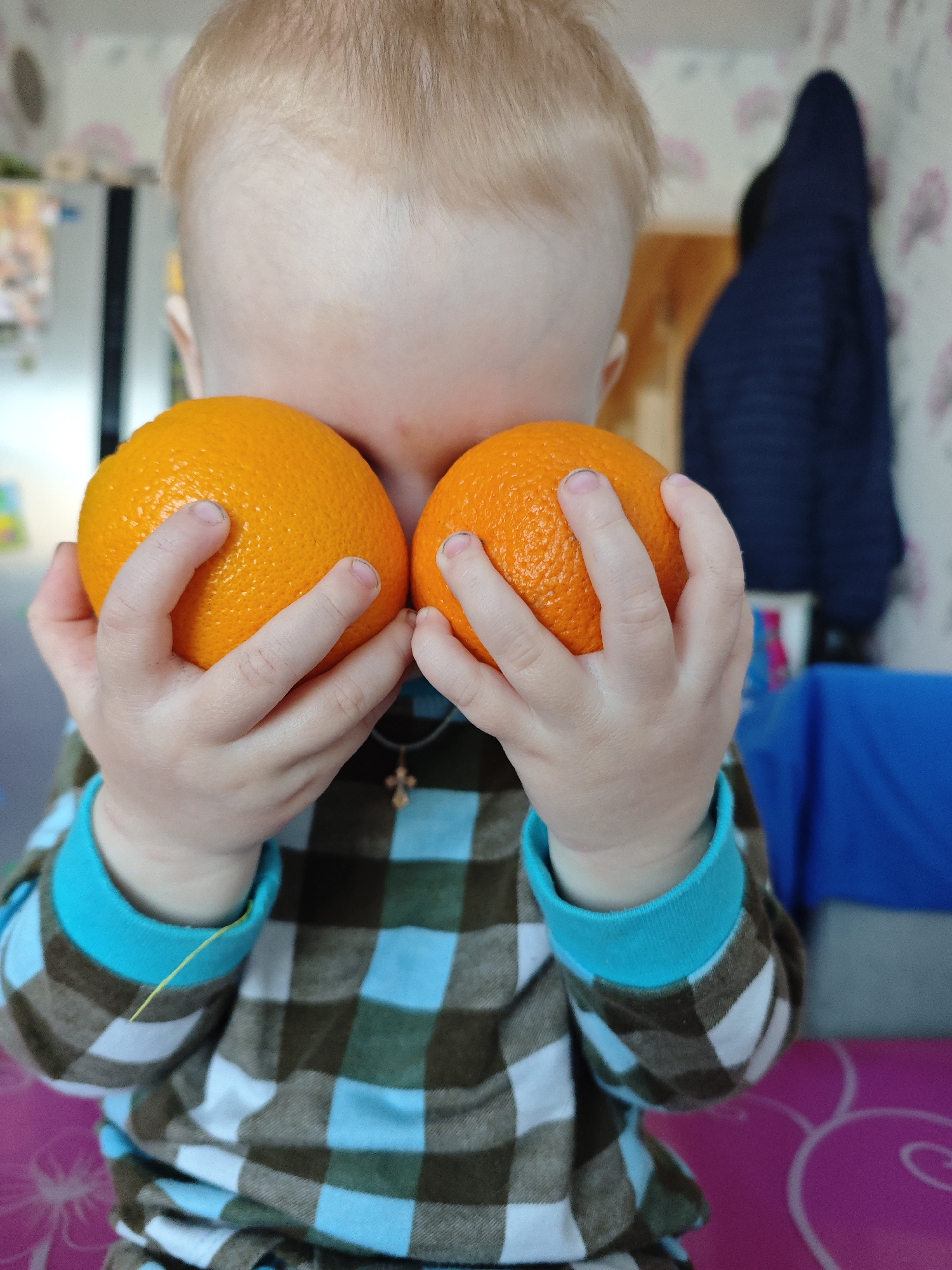 An indoor, eye-level shot captures a young child playfully holding two bright orange oranges, effectively covering most of their face. Only the crown of the child's head, with light blonde hair, is clearly visible above the fruit. Their small hands grip the oranges, with slightly dark fingertips visible.
The child is wearing a long-sleeved shirt featuring a brown, white, and light blue plaid pattern, with solid light blue cuffs. A small dark cross pendant on a string is visible around their neck, nestled between the oranges. A thin yellow thread or string hangs near the child's left wrist.
The background is softly blurred, indicating a shallow depth of field focused on the child and the fruit. To the left, indistinct shapes suggest a domestic interior, possibly kitchen elements like a refrigerator or cabinets. To the right, a dark blue quilted jacket or coat hangs on a hook against a light-colored wall adorned with a subtle floral or patterned wallpaper. In the bottom right foreground, a vibrant pink surface with a white swirling design, possibly a mat or part of furniture, adds a contrasting color to the scene.
The image conveys a moment of innocent playfulness or curiosity as the child interacts with the fruit. The lighting is bright and diffused, suggesting a daytime indoor setting. There is no visible text in the image. The scene depicts a cozy home environment, aligning with the provided location context of Markovskii, Russia.