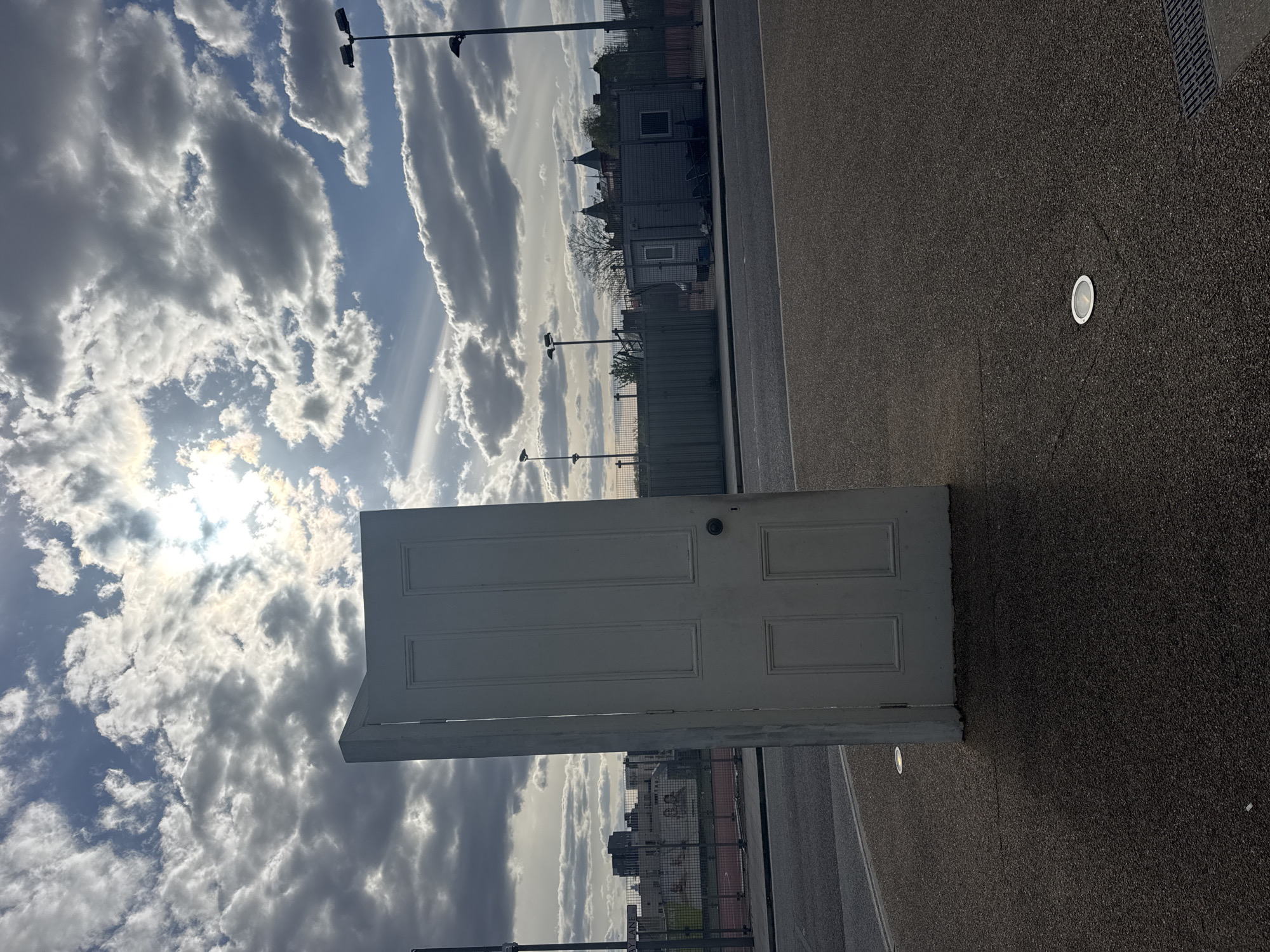 A photograph captures a white, unpainted wooden door laid horizontally on a dark asphalt or concrete surface. The door features recessed rectangular panels. The background is split between a cityscape and a bright, cloudy sky with the sun shining through. The setting appears to be London, United Kingdom, based on the architecture in the background. There are no people visible.