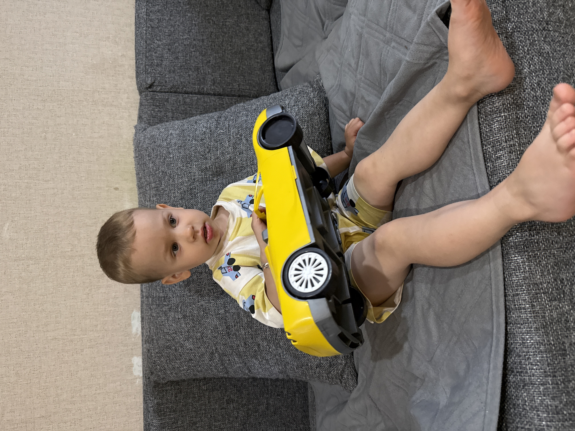 An image taken in Шебунино, Russia, shows a young child, likely a toddler, sitting or reclining on a dark grey fabric sofa indoors. The child, with light brown hair and fair skin, is looking directly at the camera with a neutral expression. He is wearing a light-colored, short-sleeved shirt with what appear to be cartoon character prints, and light-colored shorts, and is barefoot.
The child is holding a prominent, large yellow toy car with black and grey details, including white-spoked wheels, in his hands. A grey textured blanket or throw covers part of the sofa and the child's legs. To the side of the sofa, a light-colored, textured carpet or rug is visible on the floor. The setting appears to be a well-lit room, possibly during the daytime. There is no visible text in the image.