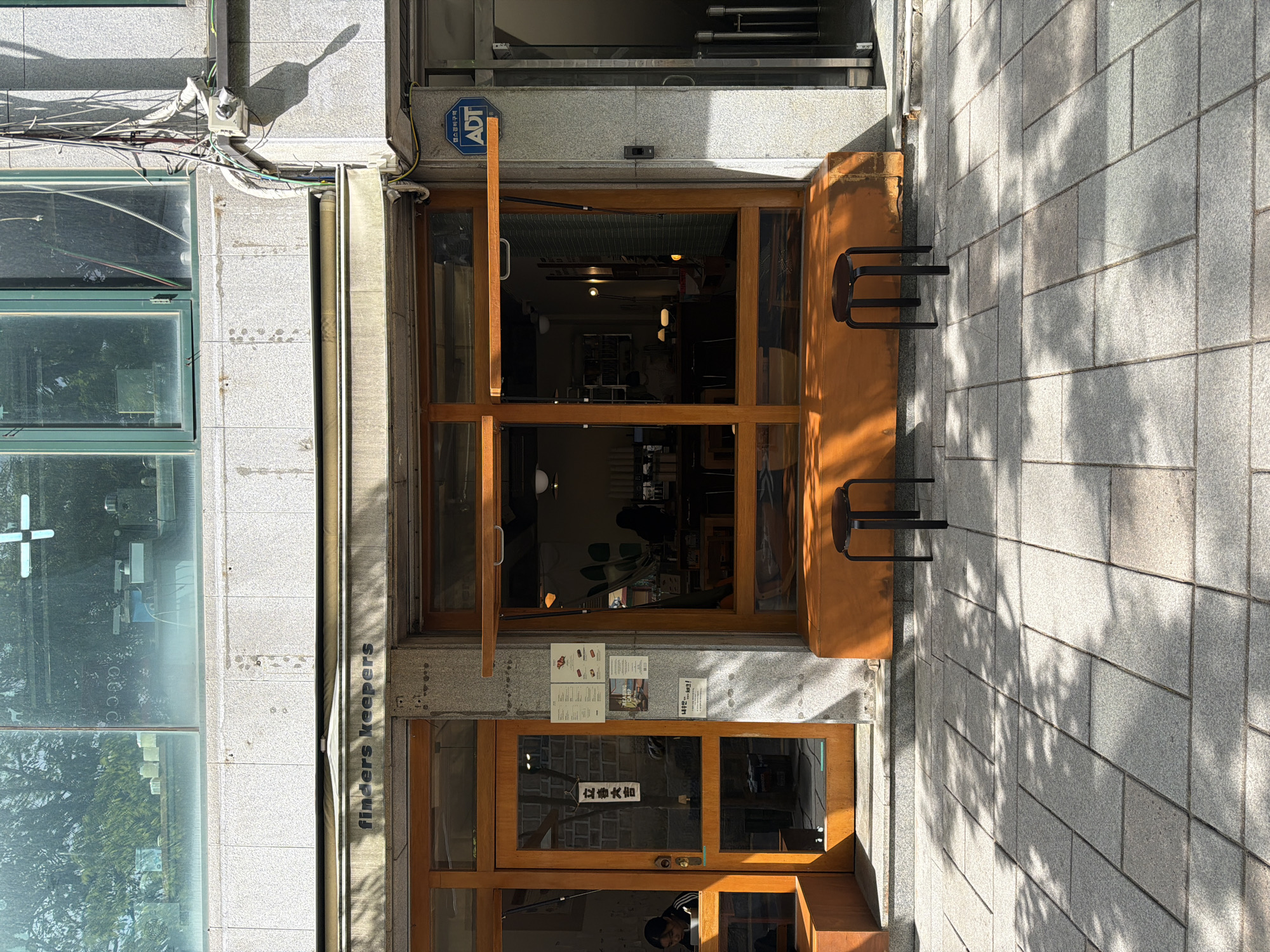 The image shows the storefront of a business called "finders keepers" in Seoul, South Korea. The front of the store has wooden-framed windows and doors. The top door is ajar, revealing the interior. Two metal stools are affixed to the right wall. A sign in the lower door has Korean text. A security system sticker is on the building above the door. The exterior of the building is light gray, and the sidewalk is paved with gray bricks. The sun casts shadows across the sidewalk.