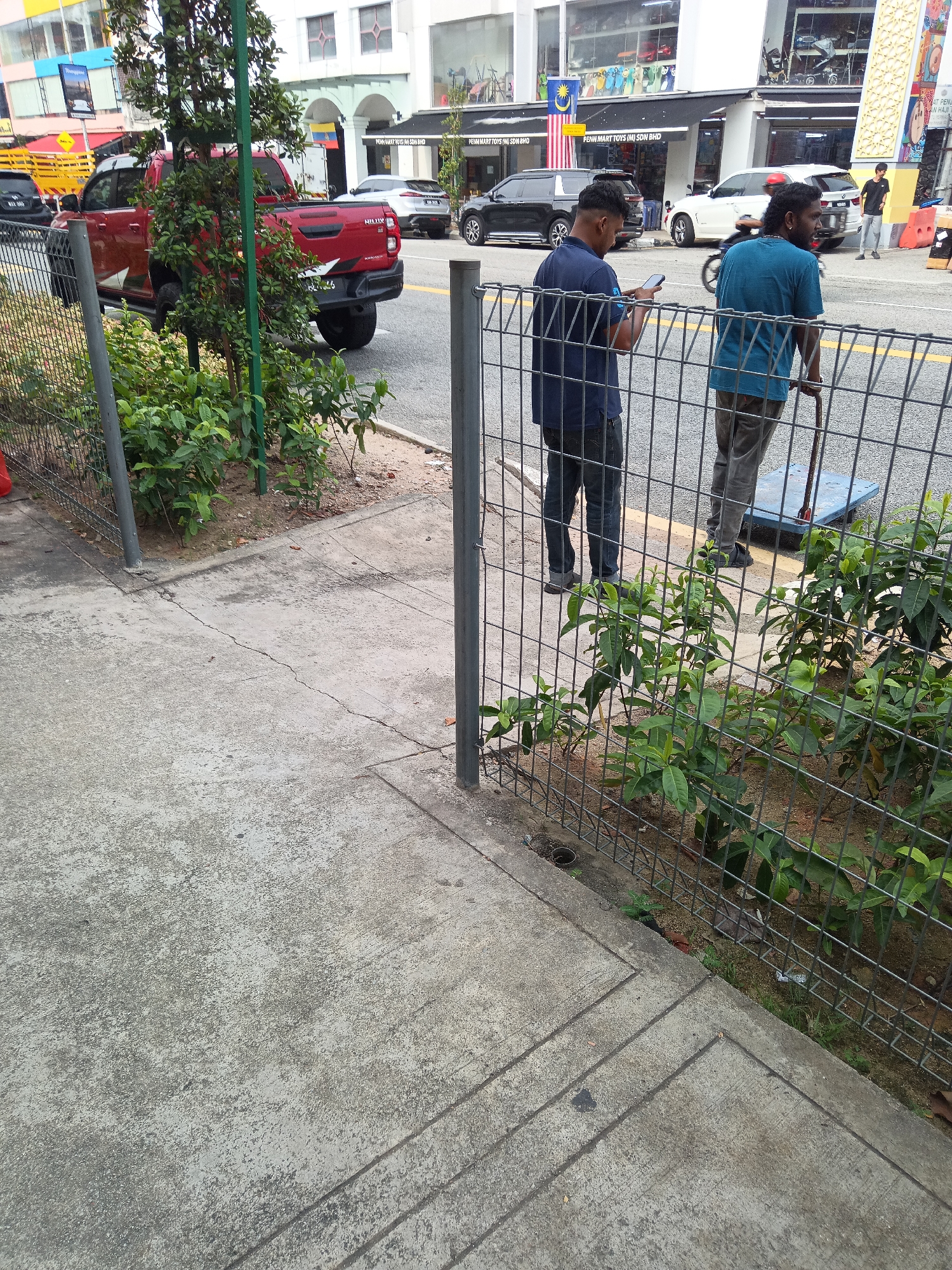 The image depicts a street scene in Kuala Lumpur, Malaysia, specifically in front of a row of shops. Two men are visible in the foreground, standing behind a wire fence. One man, wearing a blue t-shirt, is looking at his phone. The other man, in a teal t-shirt, appears to be holding onto the handle of a blue trolley. Beyond the fence, a red pickup truck is parked on the street, and several cars are also visible, including a white SUV and a black SUV. The buildings in the background are commercial establishments, with shop signs indicating businesses like "PENN MART TOYS (M) SDN BHD". A Malaysian flag is displayed prominently on one of the buildings. The environment is a typical urban street with a sidewalk and a road. The overall impression is a snapshot of daily life in a commercial district.