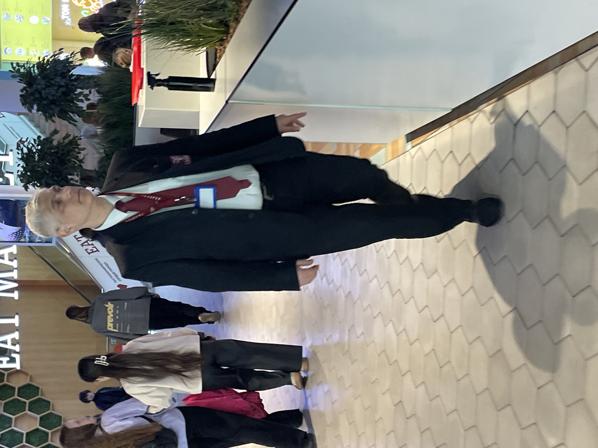 A middle-aged man in a dark suit with a red tie and a name badge is standing in a well-lit indoor space. He is gesturing with his right hand. Behind him, there are plants, a counter, and people in the background. In the foreground, several people are walking by, and a sign with the words "EAT MA..." is partially visible. The floor has a patterned tile design. The photo seems to be taken at an event in a public space. While the location shares similar visual elements with the style of the city of Saint Petersburg, Russia, the location cannot be accurately determined.