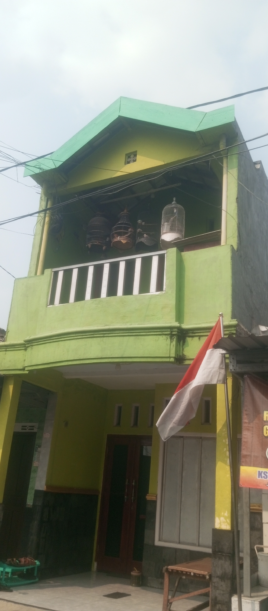 In Kediri City, Indonesia, a two-story building with a bright green exterior is captured from a low angle. The building features a prominent green roof with a sharp, modern design. The upper level has a balcony with white railings, and several bird cages are suspended from the ceiling of the covered area above the balcony, containing what appear to be birds. A white and red Indonesian flag is planted on a pole to the right of the building, partially obscuring a banner with text in Indonesian. The lower level has a yellow facade with dark gray lower walls, and a dark wooden door is visible. The ground in front of the building appears to be tiled, with some outdoor furniture and a green plastic shelf visible. Overhead, electrical wires crisscross against a light, overcast sky.