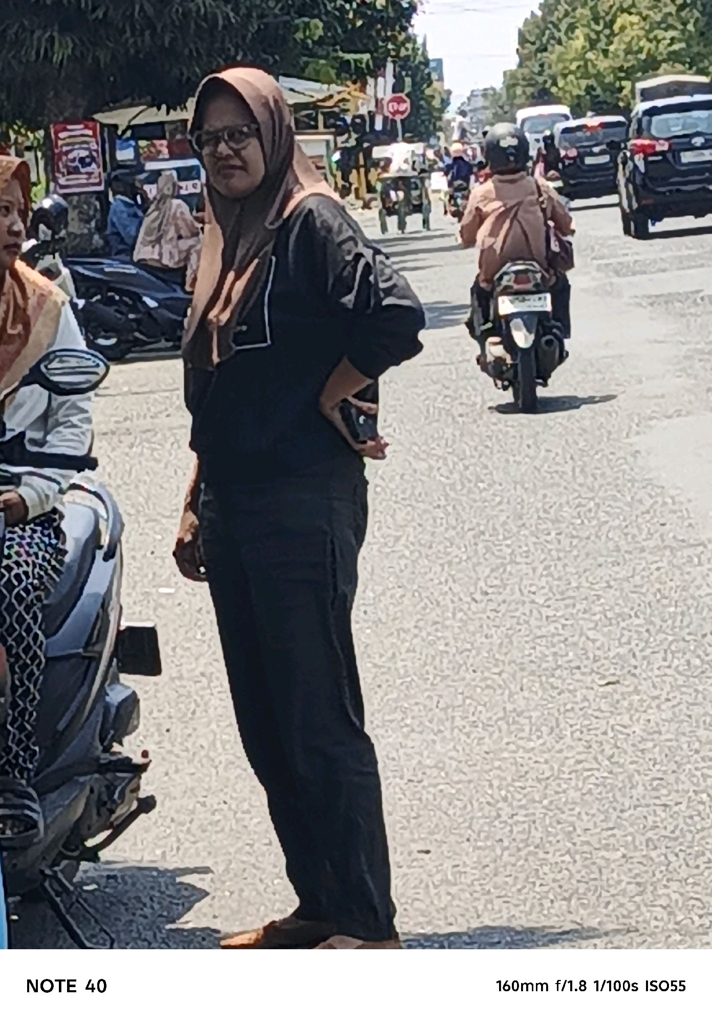 The image captures a street scene in Cilacap, Indonesia, characterized by a paved road, parked motorcycles, and busy traffic. In the foreground, a woman wearing glasses and a hijab stands facing away from the viewer, with her hand on her hip. She is dressed in a black long-sleeved shirt and dark pants. To her left, another person, also partially visible, is seated on a motorcycle. In the background, traffic includes cars and several motorcycles with riders, some of whom are also wearing hijabs. The environment appears to be an urban setting with trees lining the sides of the road, and a bright, sunny sky. The overall atmosphere suggests a typical day in a bustling Indonesian city.