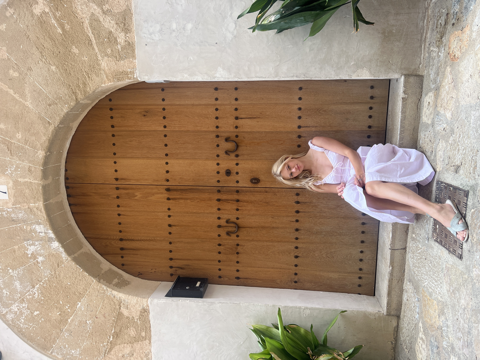 A young, blonde woman in a white sundress sits on the stone steps of an arched wooden doorway in the town of Pollença, Spain. The doorway, made of vertical wooden planks and studded with dark metal accents, features a large, U-shaped metal handle and a keyhole. Above the doorway, a rough-hewn stone archway is visible, with a numeral '7' appearing at the top. To the left of the door, a black mailbox is mounted on a white plastered wall, and a lush green plant with broad leaves grows nearby. To the right, another green plant with similar leaves is partially visible. The ground in front of the doorway is paved with textured stone, and a metal grate is set into the paving stones near the woman's feet. The woman is looking towards the camera with a slight smile, her legs crossed, and her hands resting on her lap. The overall impression is one of a serene and picturesque scene in a historic Mediterranean town.
