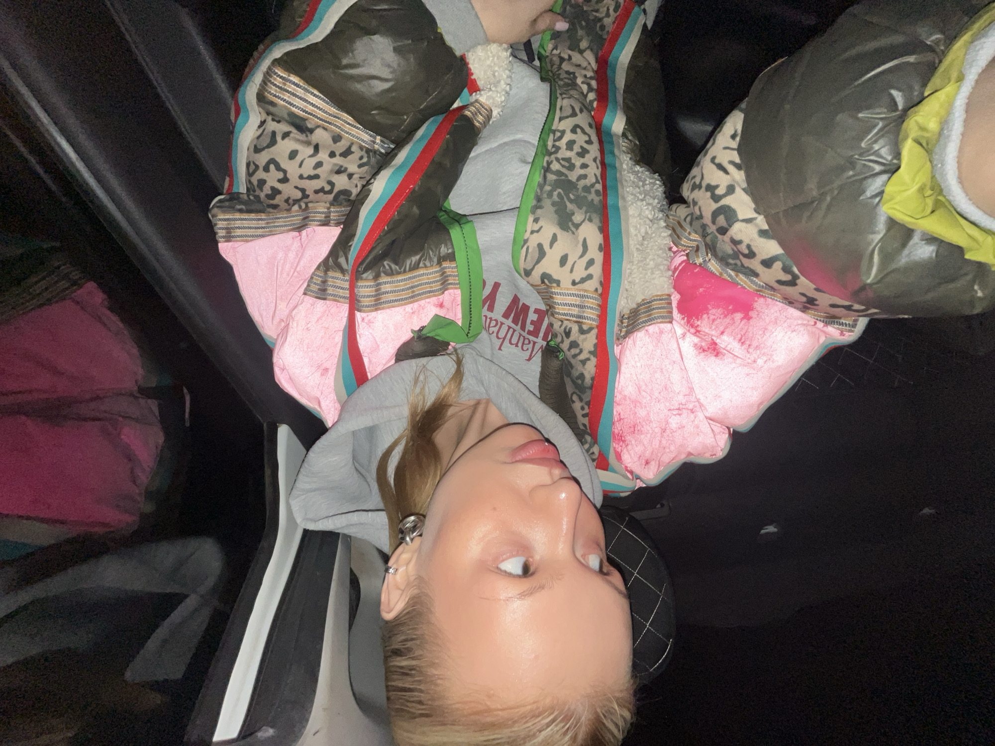 A person lies in what appears to be the back seat of a car. The person has blonde hair and is wearing a grey hooded sweatshirt with "Manhattan New York" printed on it. They are also wearing a multicolored jacket with leopard print, pink, and other patterned elements. The back seat of the car is black, and there is a pink item on the left side of the image. The location is unknown, and there is no evidence this picture was taken in Chaikovsky, Russia.