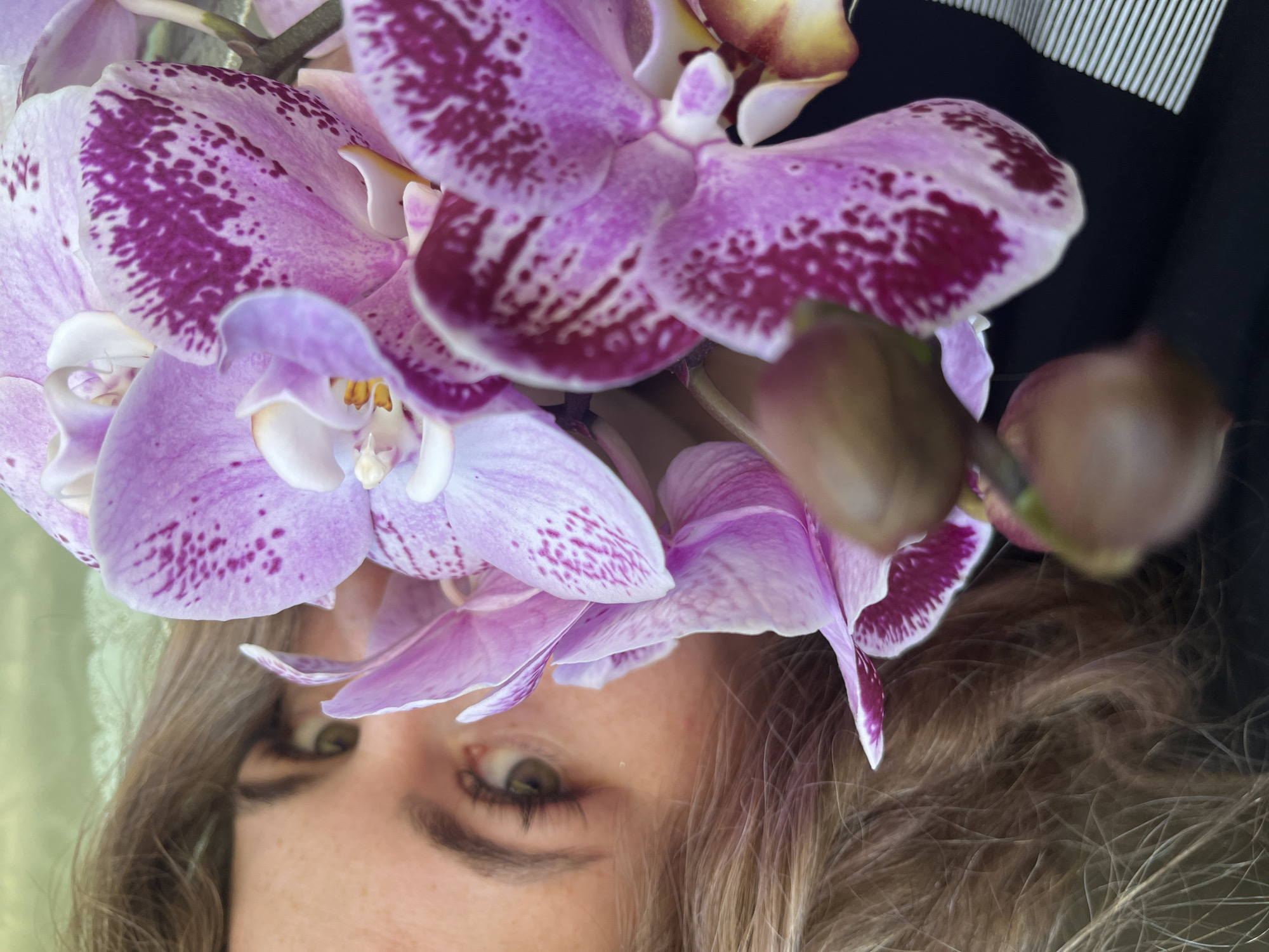 The image shows a close-up, tilted shot of a person's face partially obscured by a cluster of orchid flowers. The person has light-colored hair and green eyes, with freckles visible on their face. The orchids are predominantly light purple with dark purple speckles and spots. Behind the orchids and person, there's a glimpse of what appears to be a dark, possibly black, background with white striped pattern. The location is not discernible from the image.