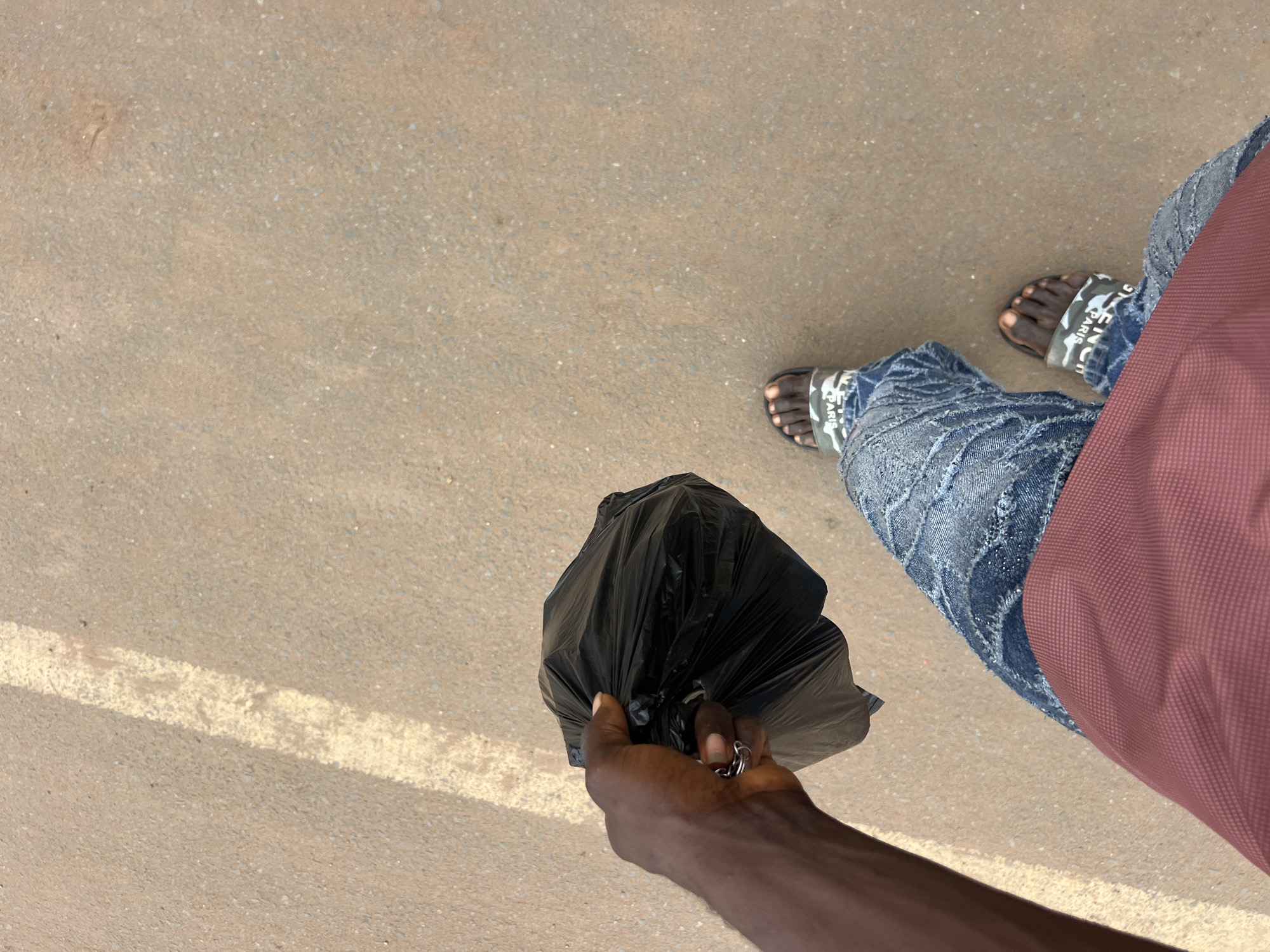 An over-the-shoulder, first-person perspective shot shows a dark-skinned hand holding a crumpled black trash bag in Nkpor, Nigeria. The hand, adorned with a silver ring, grips the bag tightly. Below, the individual is wearing distressed blue jeans with frayed patches and a maroon t-shirt. Their feet are clad in patterned sandals with the word "PARIS" visible on one strap. The ground is a textured, light brown asphalt with a faint white line running diagonally across the frame.