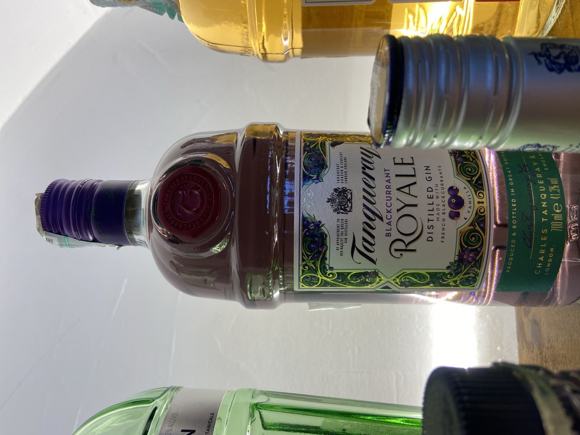 The image shows a Tanqueray Blackcurrant Royale gin bottle. The bottle has a purple top and a red wax seal. The label is green and white with purple blackcurrant designs. The label reads "Tanqueray BLACKCURRANT ROYALE DISTILLED GIN MADE WITH FRENCH BLACKCURRANTS & VANILLA" and also "CHARLES TANQUERAY & LONDON." The bottle is 700 ml and 41.3% alcohol by volume. The image also shows parts of other bottles above and below the Tanqueray bottle. The location is not mentioned in the available information.