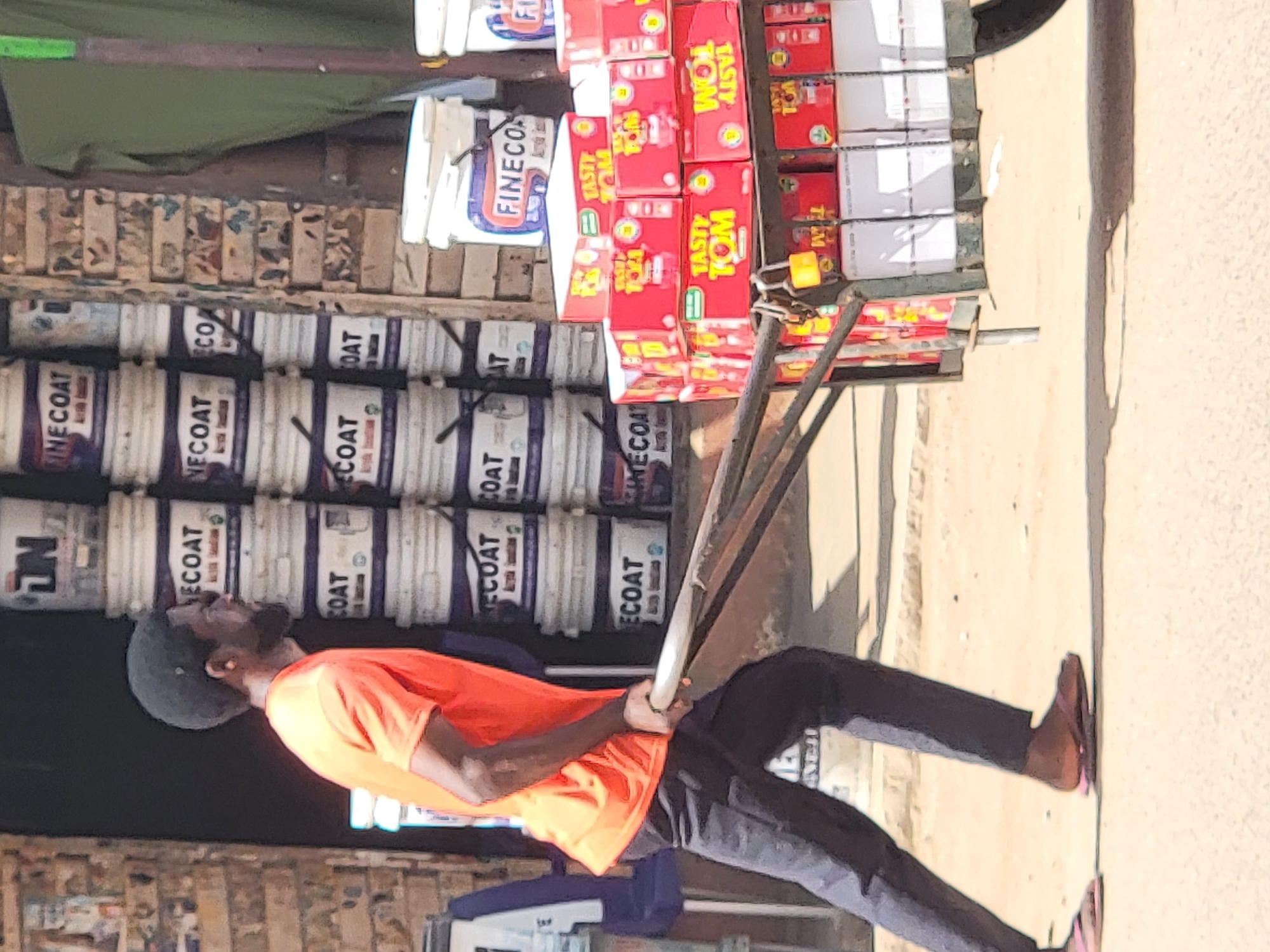 A man pulls a handcart loaded with goods in Maiburiji, Nigeria. The handcart is carrying stacks of "Tasty Tom" tomato paste and containers of "Finecoat" paint. The man is wearing an orange shirt, dark pants, a dark cap, and sandals. The background shows a shop or store with more paint containers and other items on display. The street surface is visible.