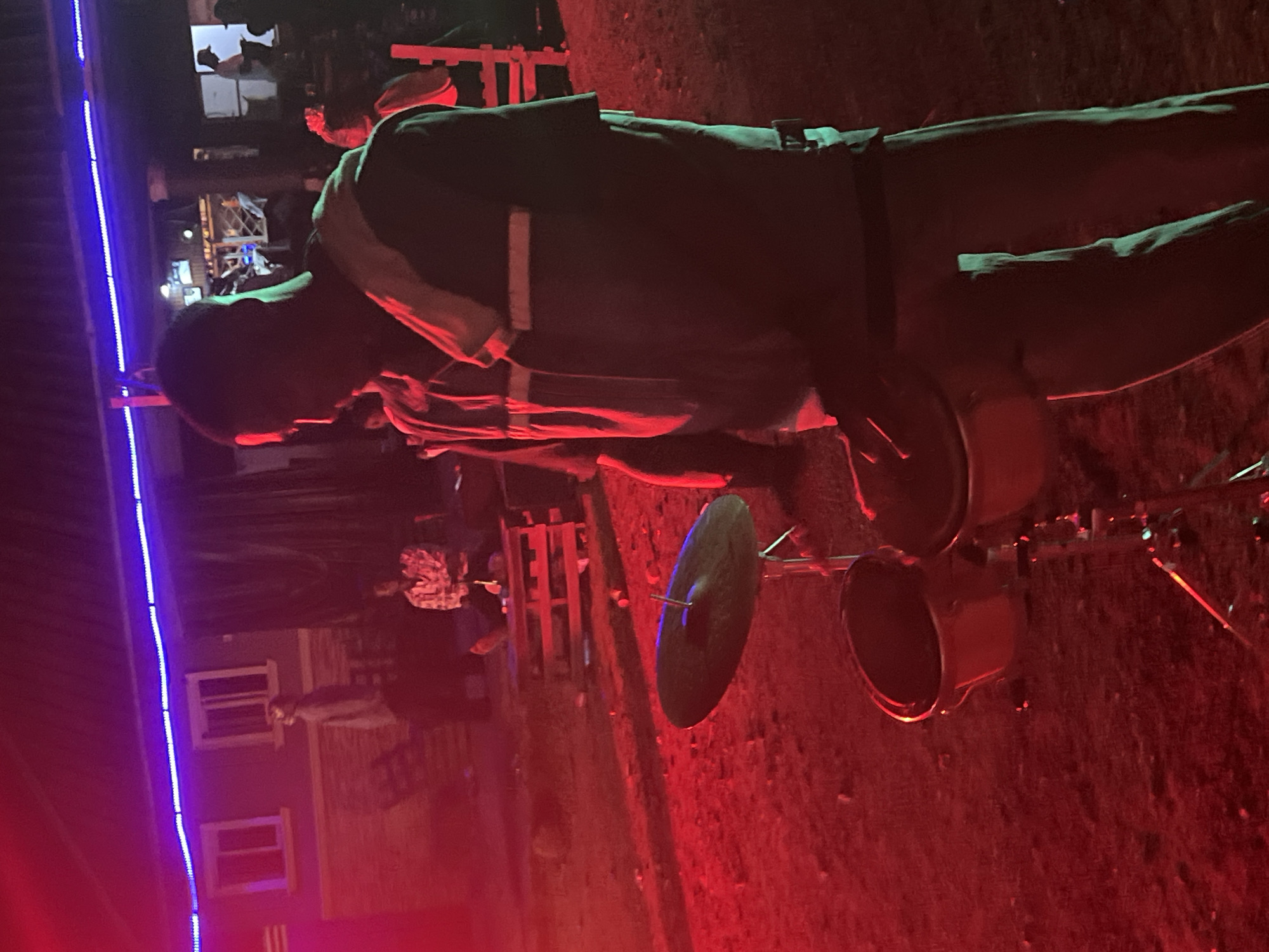 In Durumi, Nigeria, a man is standing behind a drum set, with his right hand on the snare drum, suggesting he is either playing or about to play. He is wearing a light-colored jacket with reflective stripes over a dark shirt, and dark pants. The drum set includes a snare drum and a cymbal. The scene is illuminated by red and blue lights, creating a vibrant, nighttime atmosphere. In the background, other individuals are visible, some seated and others standing, implying a gathering or event. The setting appears to be an outdoor space with a building in the background.