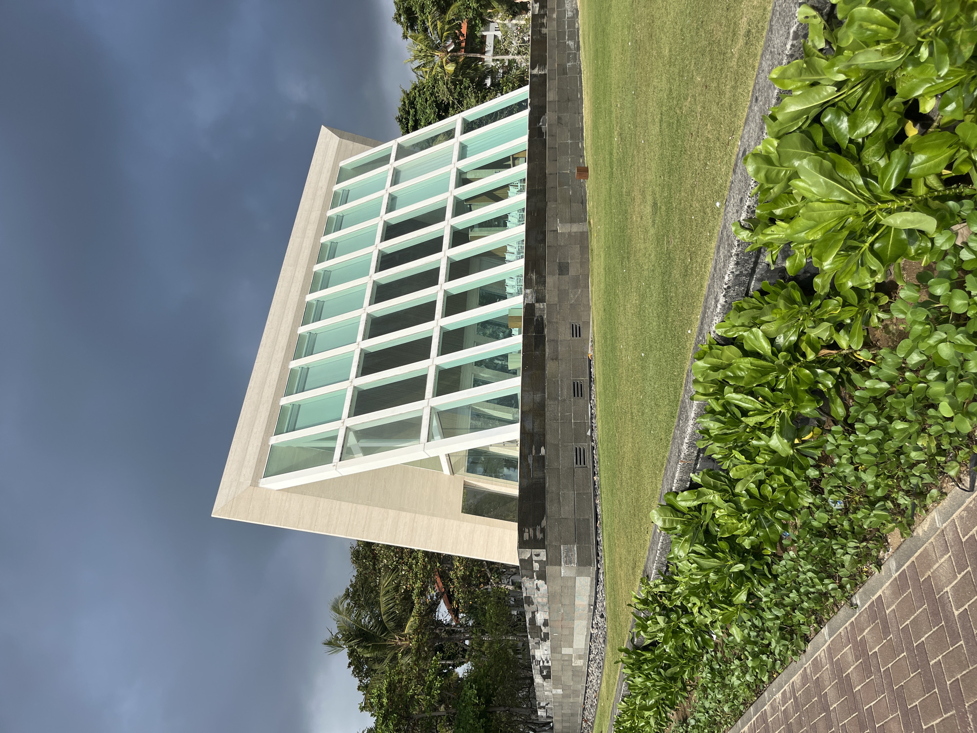 A modern glass-fronted chapel with a distinctive triangular roof stands on a grassy area in Kuta Selatan, Indonesia. The building features a light-colored, textured exterior and large, vertically paneled glass windows. It is elevated on a dark stone base. Surrounding the chapel is a manicured lawn, bordered by lush green foliage and a brick pathway in the foreground. In the background, palm trees and other vegetation are visible, along with parts of other buildings. The sky is overcast and dark, suggesting potential rain. There are no people visible in the image.