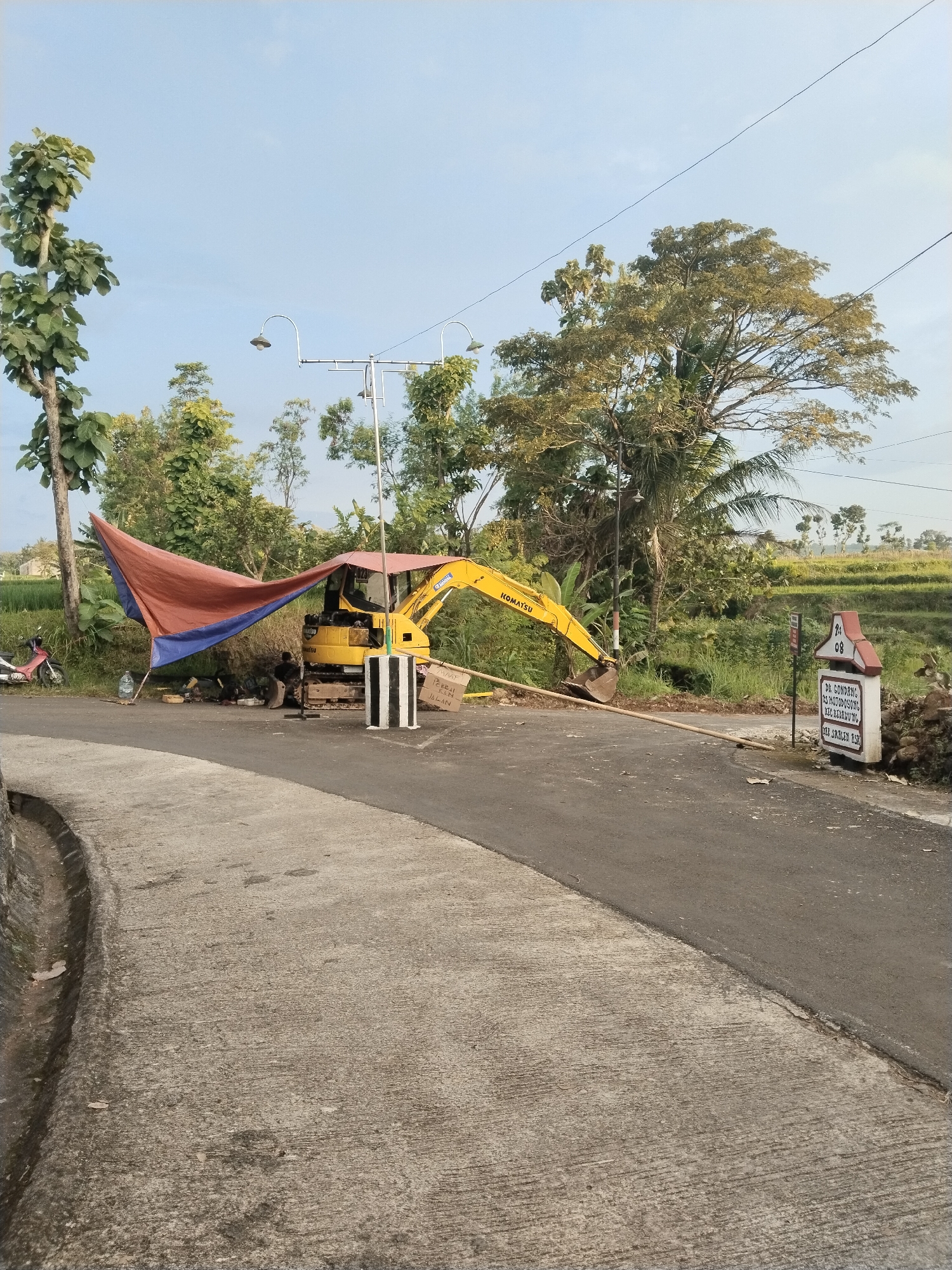 The image shows a road construction scene in an area identified by a sign as "Ds. Gondang, Ds. Hotododoyong, Kec. Kedawung, Kab. Sregen Asr." A yellow Komatsu excavator is parked on the side of the road, partially covered by a makeshift tarp shelter supported by poles. There is a person or object obscured under the tarp. The road is divided into two distinct surfaces - a rough concrete section and a smooth asphalt section. A black and white striped barrier stands next to the excavator. Nearby are a pole with several curved streetlights, a sign with the text "PERBAIKAN JALAN" ("Road Repair" in Indonesian), trees, and what appears to be a rice paddy. A motorcycle is parked in the background.