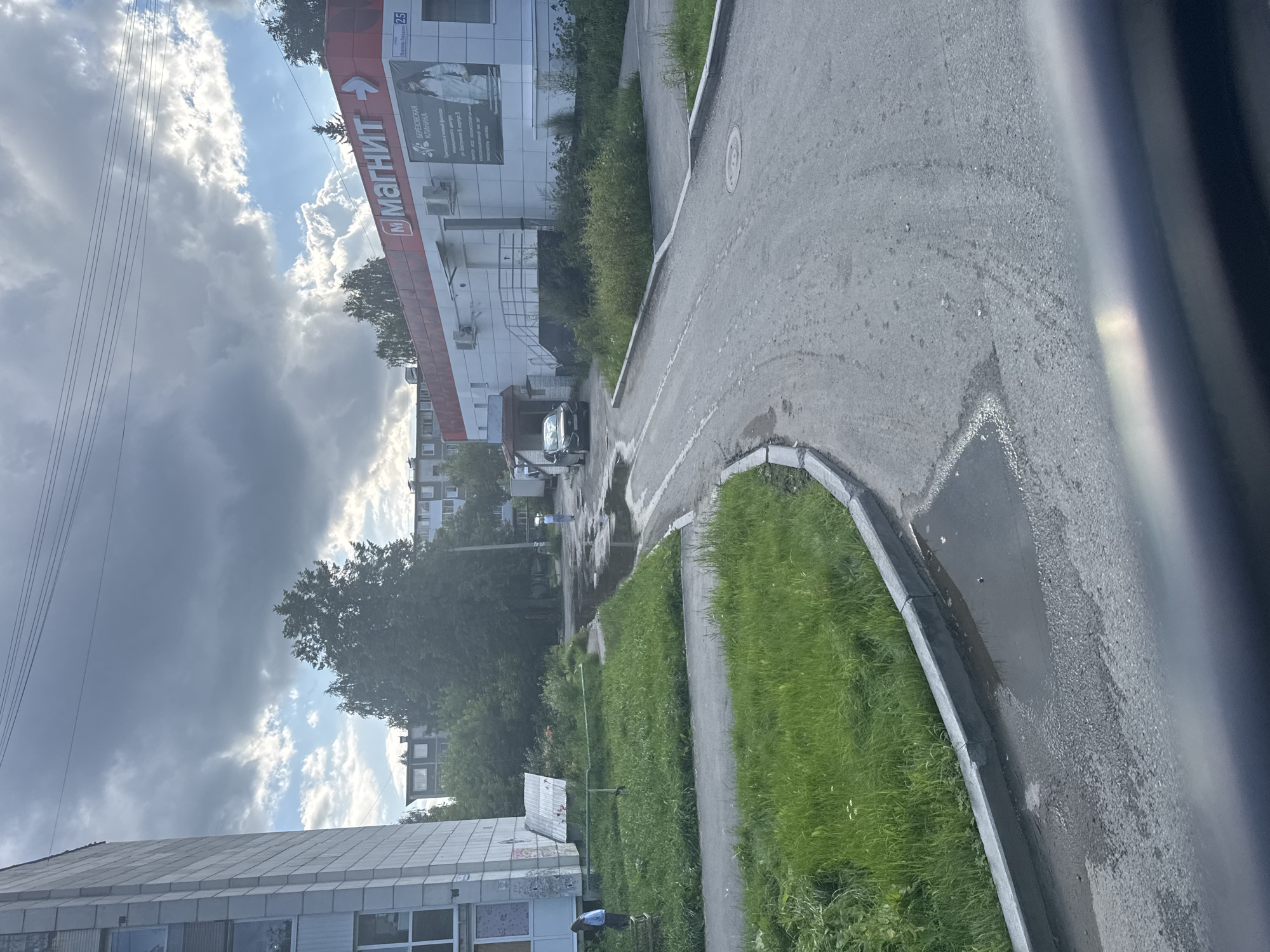 The image shows a view of a residential area in Beryozovsky, Russia, with a multi-story building on the left and a commercial building with a "Magnit" grocery store sign on the right. A dark-colored car is parked on the asphalt road in front of the commercial building, which also has an advertisement for "Beryozovskaya Klinika." Several puddles are visible on the road and adjacent grassy areas, suggesting recent rain. Green grass and trees are present throughout the scene. The sky is partly cloudy with both dark and light clouds. There are two individuals visible; one person, wearing a light blue top, is standing near the multi-story building, and another person, wearing a white top and dark pants, is walking on the road in the background. Overhead power lines are visible across the top of the frame.