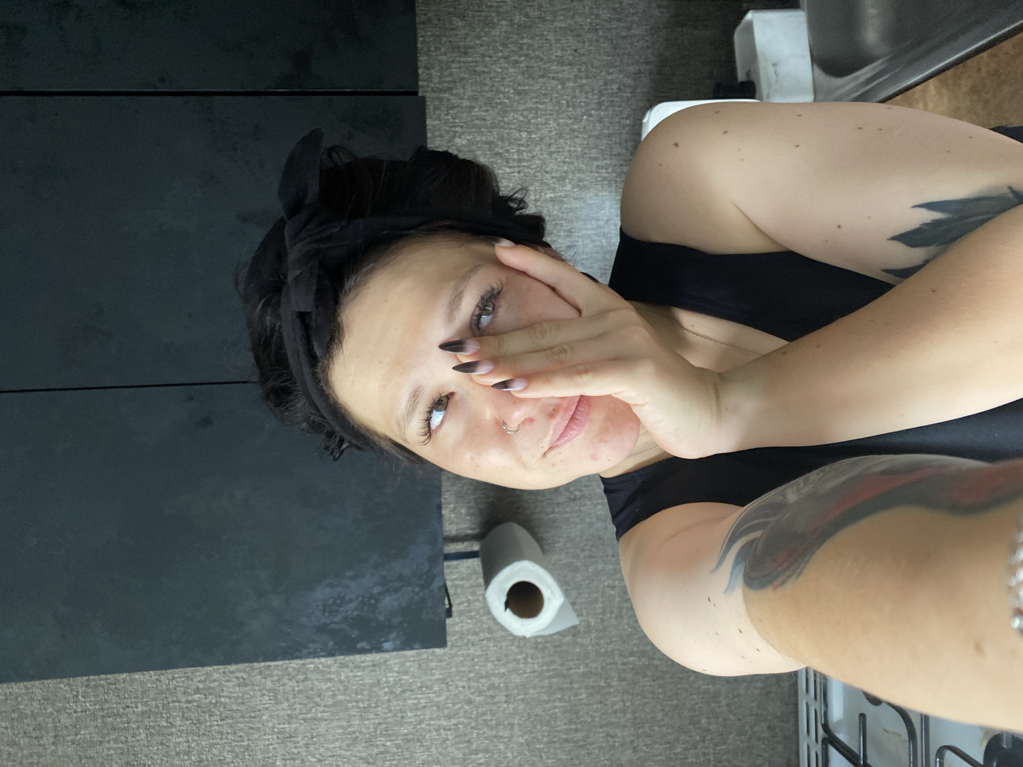 A woman with dark hair tied up with a black bandana is taking a selfie in a kitchen. She is wearing a black tank top. Her left hand is covering the lower half of her face. She has a nose ring and her fingernails are painted black and white. There is a roll of toilet paper on the wall behind her, and a stove is visible in the lower left corner of the image. The environment appears to be an indoor kitchen with grey textured walls and dark cabinets.