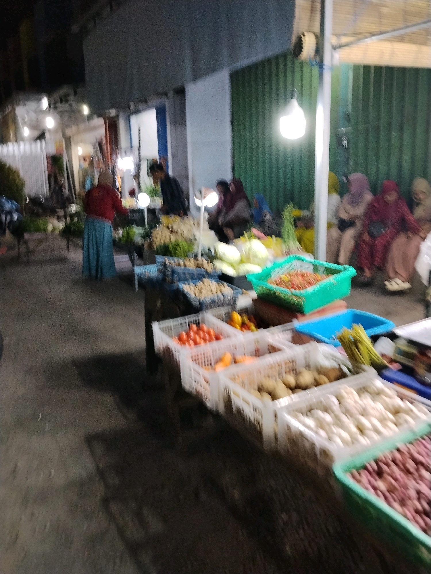 The image depicts a dimly lit outdoor market scene, likely in Panggungan, Indonesia, at night. Several individuals are present, some engaged in activities related to selling or purchasing goods. In the foreground, a variety of produce is displayed in plastic crates. These include red tomatoes, onions with purplish-red skins, and what appear to be white root vegetables or bulbs. Behind these, more produce is visible, including potatoes and what seems to be a bundle of green onions or leeks. Further back, in the middle ground, a person in a red top and a blue skirt is standing near a stall. To the right, a group of women wearing headscarves are seated. The market is illuminated by overhead lights. The overall impression is of a bustling local market at the close of day.