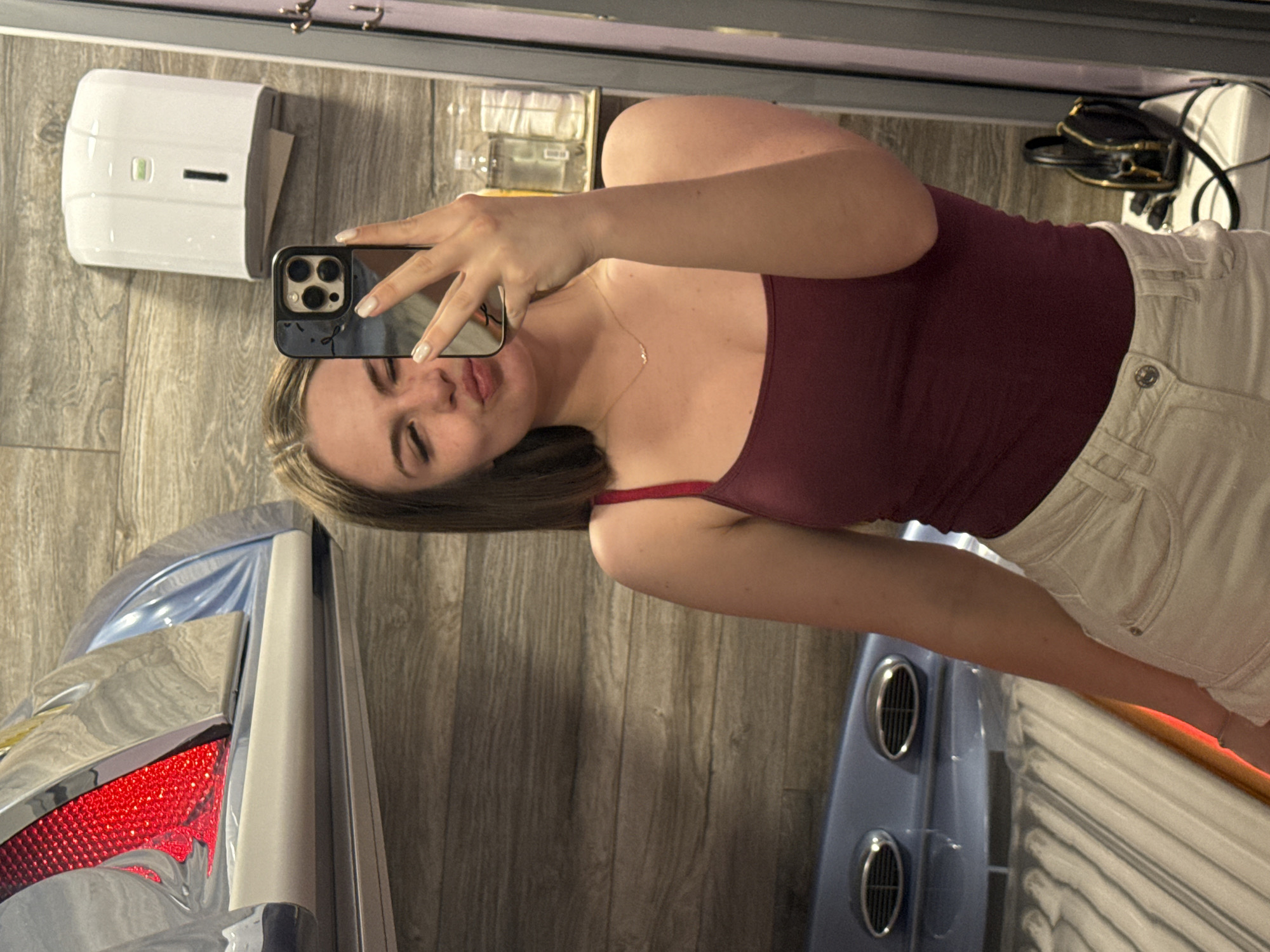 The image depicts a selfie taken in what appears to be an indoor tanning salon. A woman is holding a smartphone, capturing her reflection in a mirror. She is wearing a maroon tank top and off-white denim shorts. She has light skin and brown hair. Visible in the background are the wooden paneled walls, a white paper towel dispenser, and what seems to be tanning bed equipment, partially visible on the left and bottom of the frame. It is not possible to ascertain the specific location in Plovdiv, Bulgaria or any other location based on the visual information in the image.