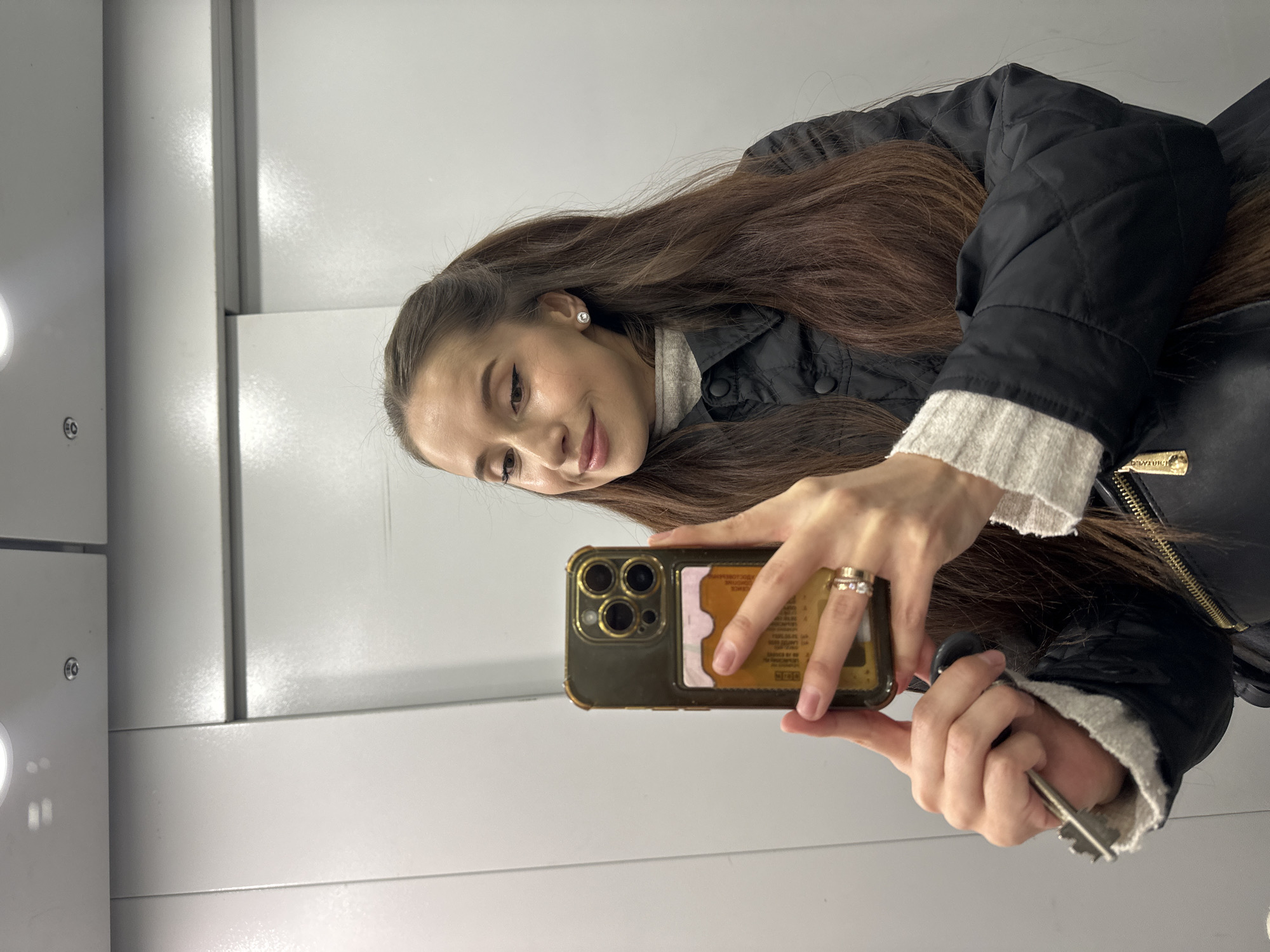 The image, rotated 90 degrees clockwise, shows a young woman taking a mirror selfie in an indoor setting, likely an elevator in Yekaterinburg, Russia. She has long, dark brown hair and a gentle smile, looking down towards her phone.
She is dressed in a light-colored, ribbed sweater underneath a black quilted jacket. The jacket features a prominent gold zipper pull with the brand name "BARBOUR" clearly visible. A small, sparkling stud earring adorns her right ear.
In her left hand, she holds a dark-gold toned smartphone, identifiable by its triple camera array, housed in a transparent case. The case has a card slot on the back, containing at least two cards, one with partially visible text including "BY ROYAL O". On her left ring finger, she wears two rings: one a plain gold band and the other a ring with a diamond or gemstone setting. Her right hand holds a single silver-colored key.
The background consists of light-colored, possibly metallic or laminated, wall panels with distinct vertical seams, characteristic of an elevator interior. A circular light fixture is partially visible on the far left of the frame, providing bright, even illumination.
