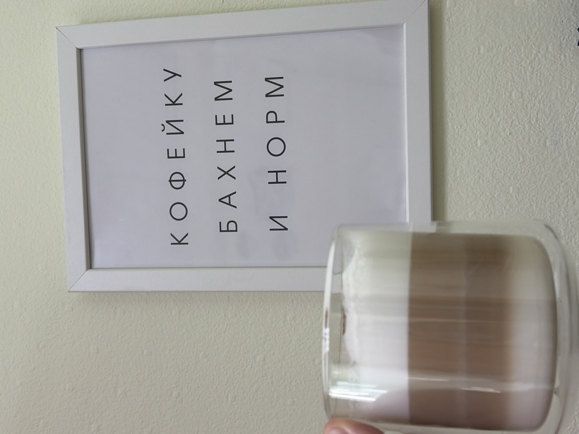 An indoor scene captured in Chaikovsky, Russia, features a framed poster on a light-colored, textured wall alongside a double-walled glass containing a layered beverage, held by a person.
The poster, oriented vertically on the left side of the frame, has a simple white border and a white background. Black sans-serif capital letters are arranged in three vertical lines. From left to right (or top to bottom if the image were rotated 90 degrees counter-clockwise), the text reads:
1. КОФЕЙКУ
2. БАХНЕМ
3. И НОРМ
This translates approximately to "A little coffee, let's have it, and it'll be fine" or "Let's drink some coffee and everything will be alright," conveying a casual, encouraging sentiment.
On the right side of the image, slightly out of focus, a transparent, double-walled glass is being held by a person's thumb, which is visible at the bottom. The glass contains a two-toned drink, likely a coffee with milk or cream, featuring a distinct darker brown layer at the bottom and a lighter, creamy white layer above it. The layering suggests a latte or similar beverage.
The wall behind the poster and drink is painted in a pale, off-white or cream color and has a visible textured, possibly stucco-like finish. The lighting appears soft and ambient, characteristic of an indoor setting. The activity involves someone holding a beverage, possibly preparing to drink it, while the framed message adds a cheerful, functional element to the background.