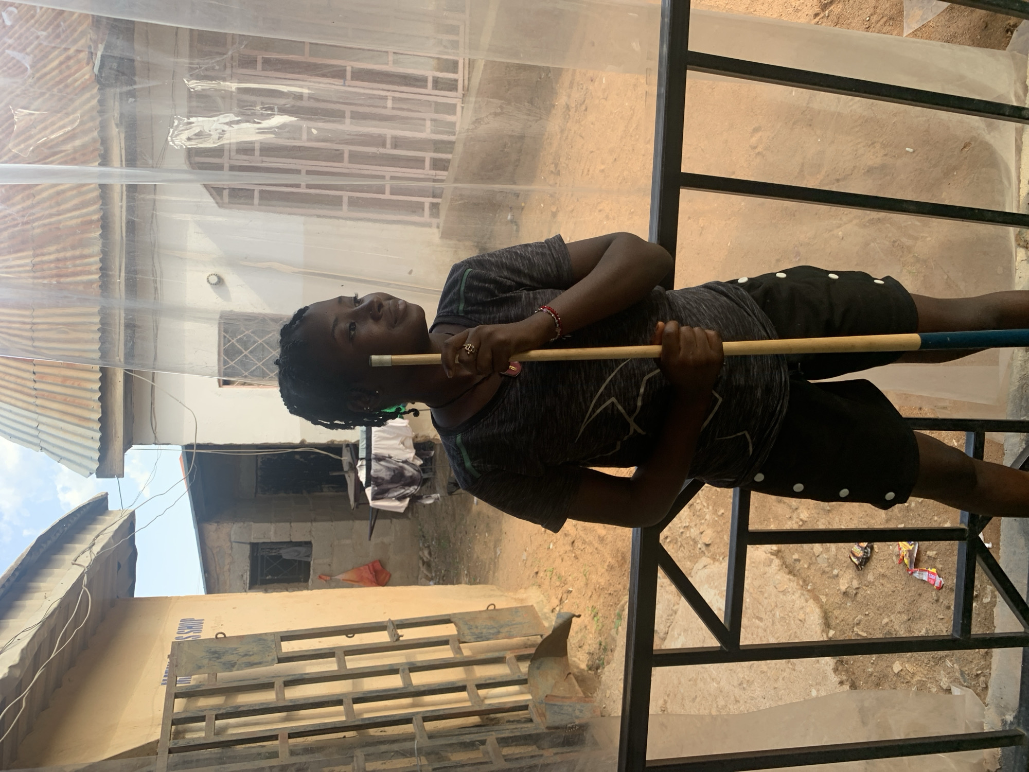 A young woman is standing in an outdoor area in Barakin Akawo, Nigeria. She holds a pool cue horizontally. The area is surrounded by black metal railings, and a clear plastic sheet is draped overhead. In the background are buildings constructed of concrete and corrugated metal. There is litter scattered on the ground.