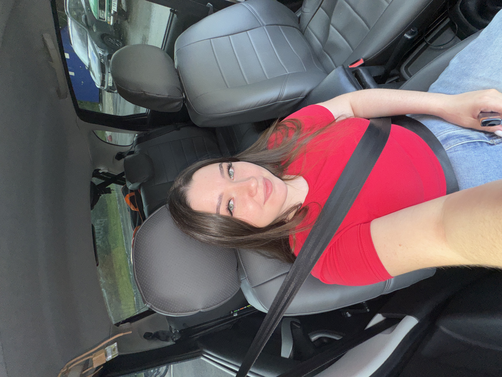 The image shows a young woman with long brown hair and blue eyes, wearing a red t-shirt and blue jeans, sitting in the passenger seat of a car. She is wearing a seatbelt. The car's interior is visible, with grey leather seats. Outside the car, a grey SUV is parked, and a portion of a green car is also visible. The setting appears to be a street or parking area in the city of Perm, Russia.