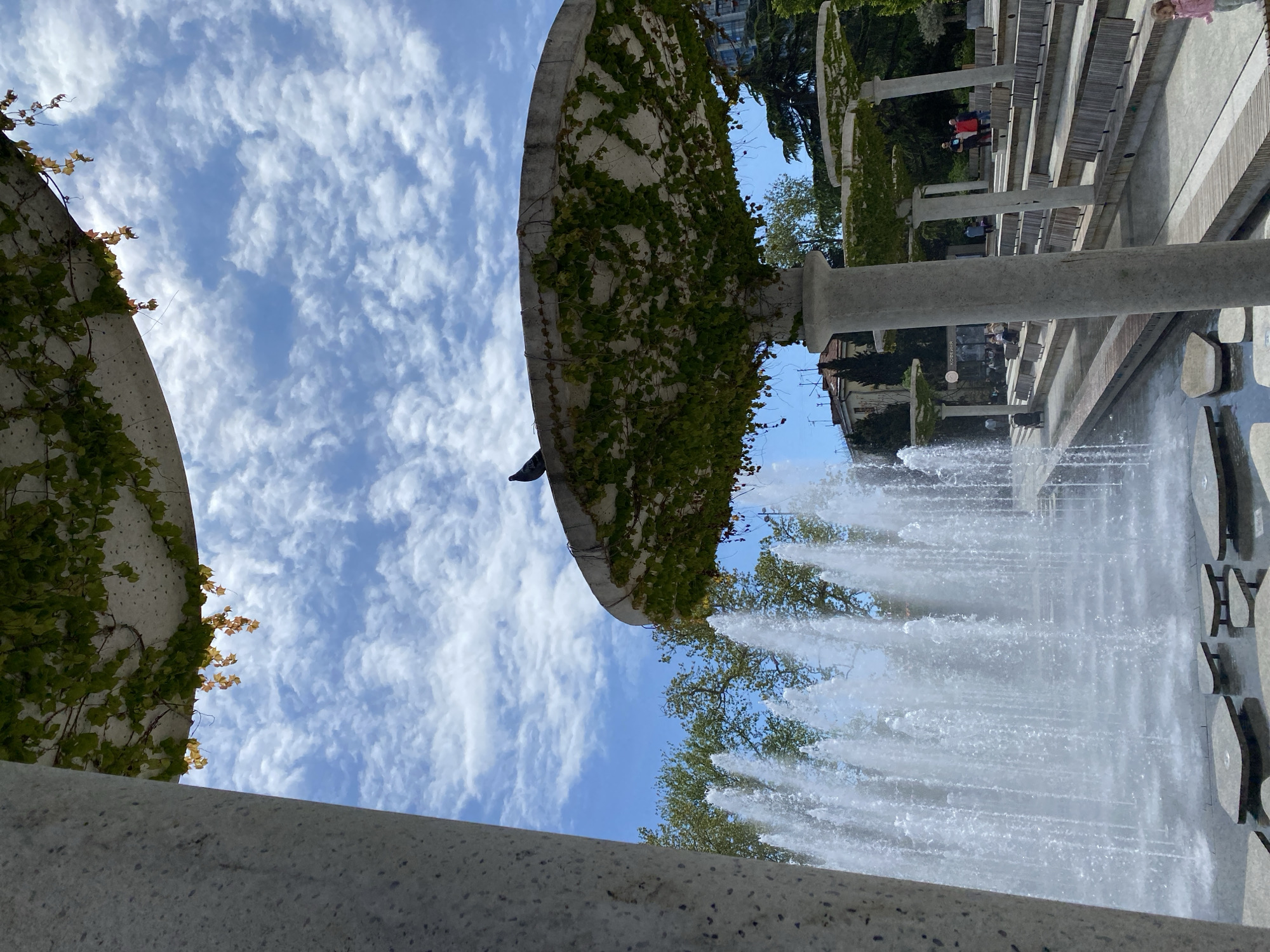 The image captures a view of a fountain in город Ялта, Ukraine, with architectural elements covered in ivy. The fountain jets water upwards, creating a dynamic scene. In the background, tiered seating and concrete structures are visible, suggesting a park or public space. People can be seen seated in the background. The sky is partly cloudy, with patches of blue visible through the clouds. The overall perspective is looking upwards, emphasizing the height of the fountain and the surrounding structures.