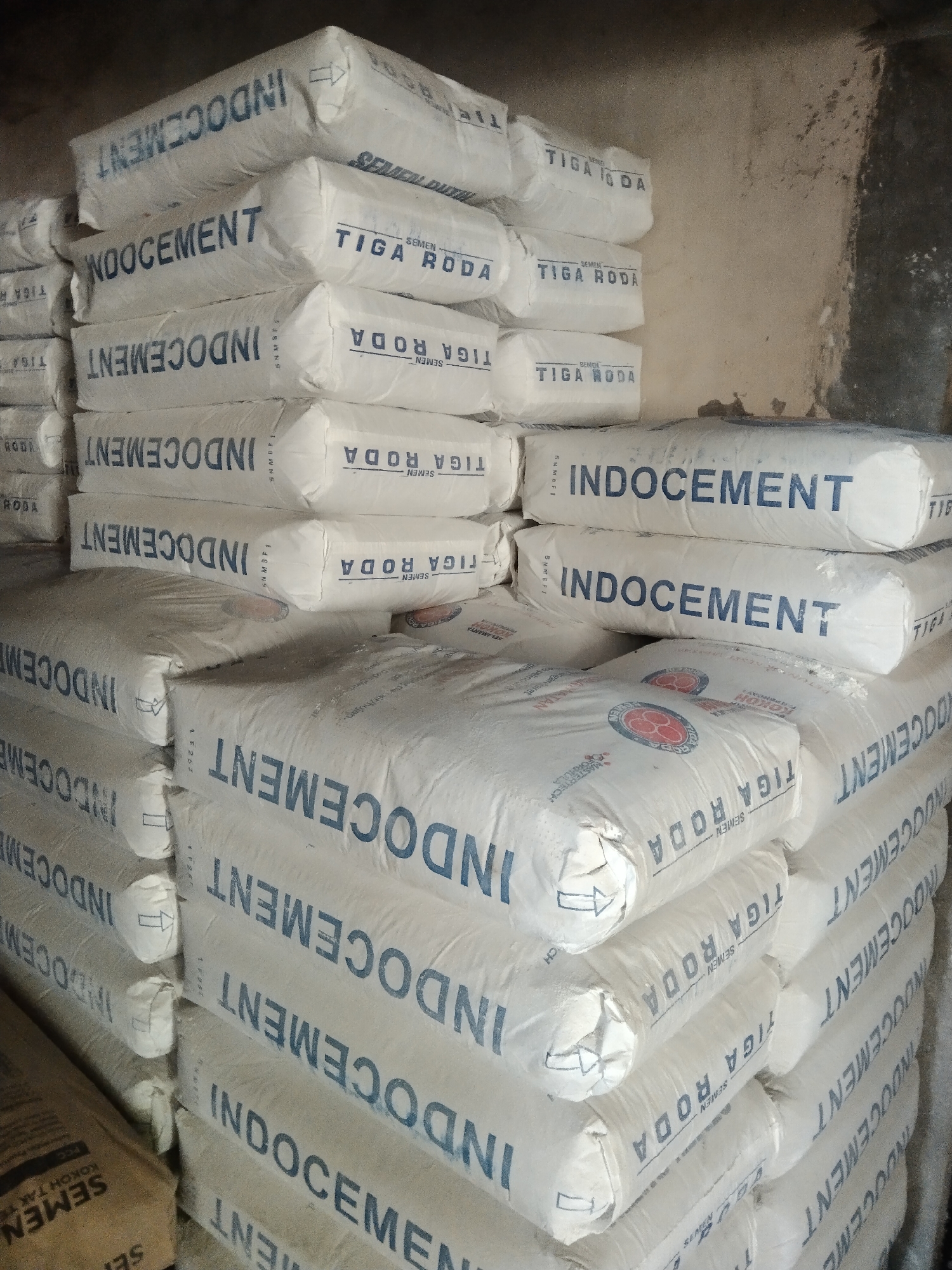 A large pile of cement bags is stacked in a dimly lit room, likely a warehouse or storage facility, in Parung, Indonesia. The bags are predominantly white with blue lettering. The most prominent branding visible is "INDOCEMENT" repeated multiple times, along with "TIGA RODA" and "SEMEN." The bags are stacked in various configurations, some forming neat rows and others appearing more haphazard. The room itself appears to be constructed of concrete blocks or similar rough materials, with shadows and dust suggesting an industrial or utilitarian setting. No people are present in the image. The lighting is uneven, with harsh shadows cast across the bags and walls, indicating a single or limited light source. The overall impression is one of a bulk storage of construction materials.