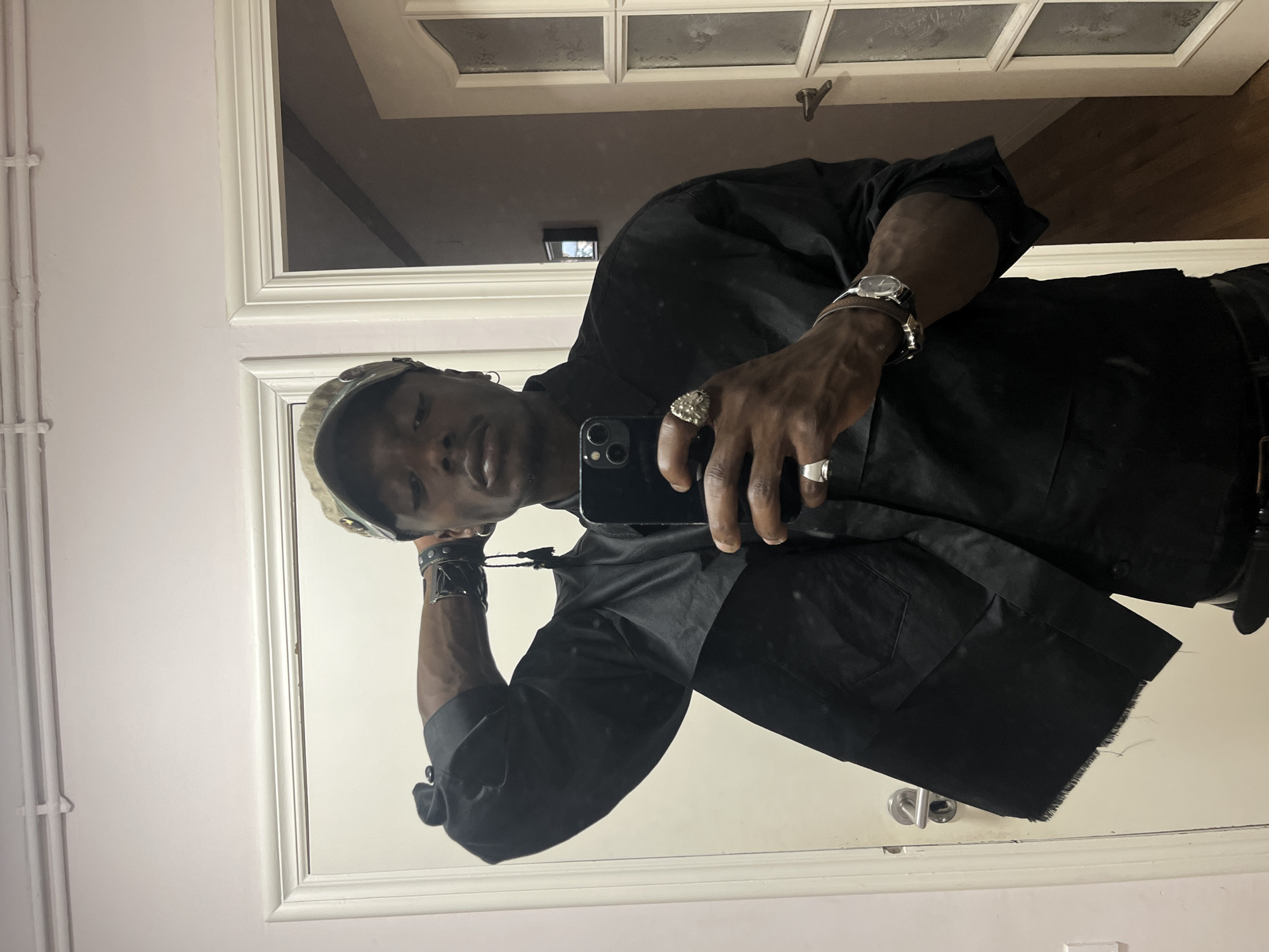 The image displays a mirror selfie, rotated 90 degrees counter-clockwise, featuring a person, likely male, captured in their reflection. The individual is shown in a tilted pose, with their head angled upwards and looking directly towards the camera (reflected in the smartphone).
The subject is wearing a dark, short-sleeved, button-up shirt or jacket. A camouflage-patterned cap or bandana covers their head. Accessories include a silver-toned watch with a dark strap on the left wrist, along with a dark, studded leather-like bracelet. Multiple rings adorn their fingers: a large, ornate silver-colored ring and a smaller plain silver ring are visible on the right hand, while a large, plain silver ring is on the left hand. They are holding a dark-colored smartphone, likely an iPhone with multiple cameras, in their right hand to take the photograph.
The setting is indoors, featuring prominent white architectural elements. White door frames, possibly from a door or window with multiple panes, are visible in the background, reflecting parts of a ceiling or an internal structure. A light-colored wall with white vertical pipes is seen on one side. A dark wooden floor is reflected at the bottom of the frame (if viewed with correct orientation). The lighting is consistent with an indoor environment.
No specific activity beyond taking the selfie is depicted. The person's expression is neutral. There are no visible texts in the image. While the location is provided as Barcelona, Spain, there are no visual cues within the image itself to confirm this.