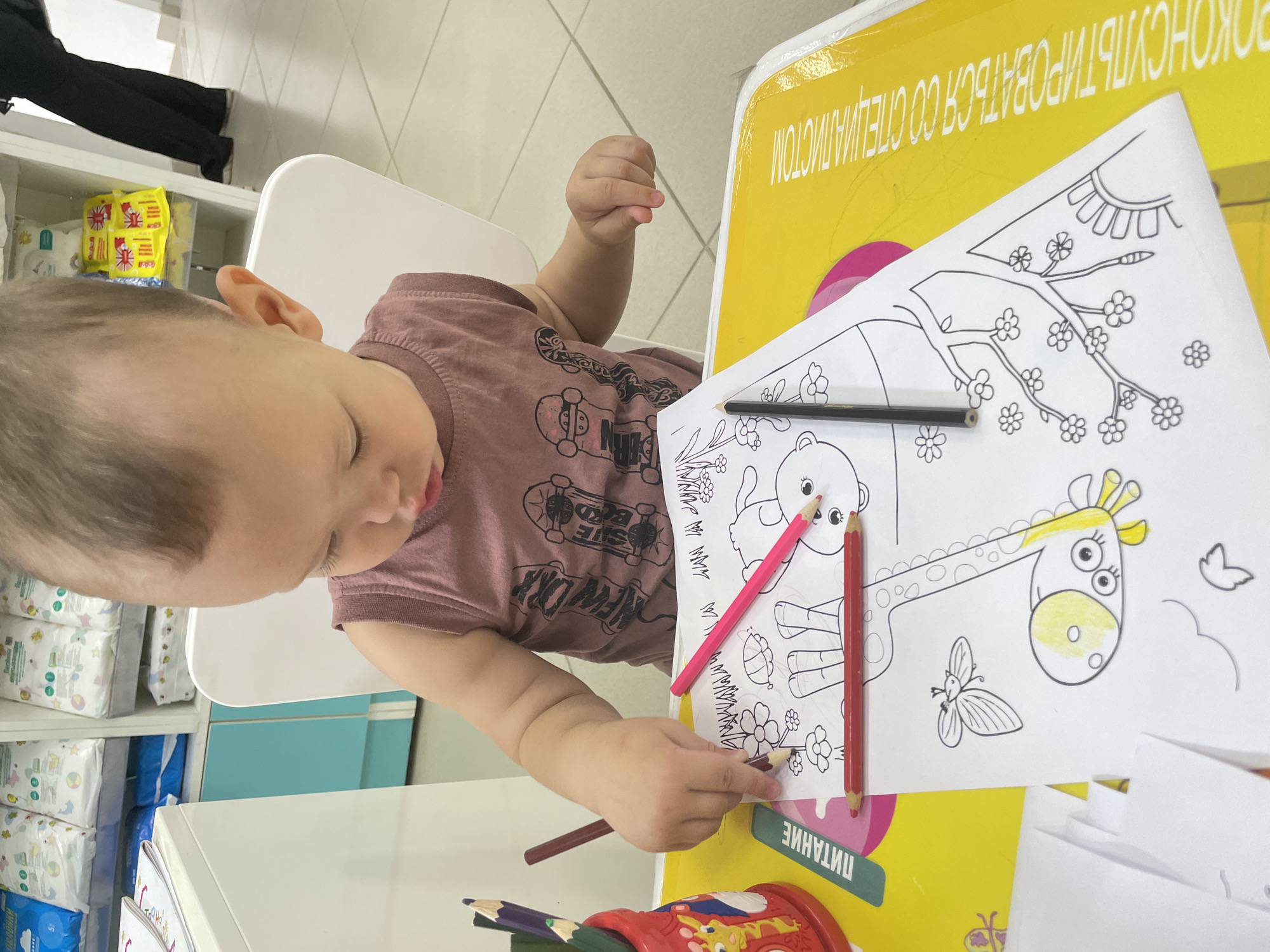 A toddler sits at a table, coloring a picture of a giraffe and flowers. The toddler is wearing a sleeveless shirt and is focused on the coloring book, holding a brown colored pencil. Various other colored pencils are scattered on the table, along with a red pencil cup filled with more pencils. A green label with Russian text "ПИТАНИЕ" is visible on the left side of the table. The table is covered with a yellow plastic mat featuring yellow and white text. In the background, shelves are stocked with packages of baby diapers and other baby care products. The scene appears to be indoors, possibly in a store or a dedicated play area, as suggested by the retail packaging in the background. The lighting is bright, indicating an indoor setting with artificial or ample natural light. The location of the image is not specified within the visual data.
