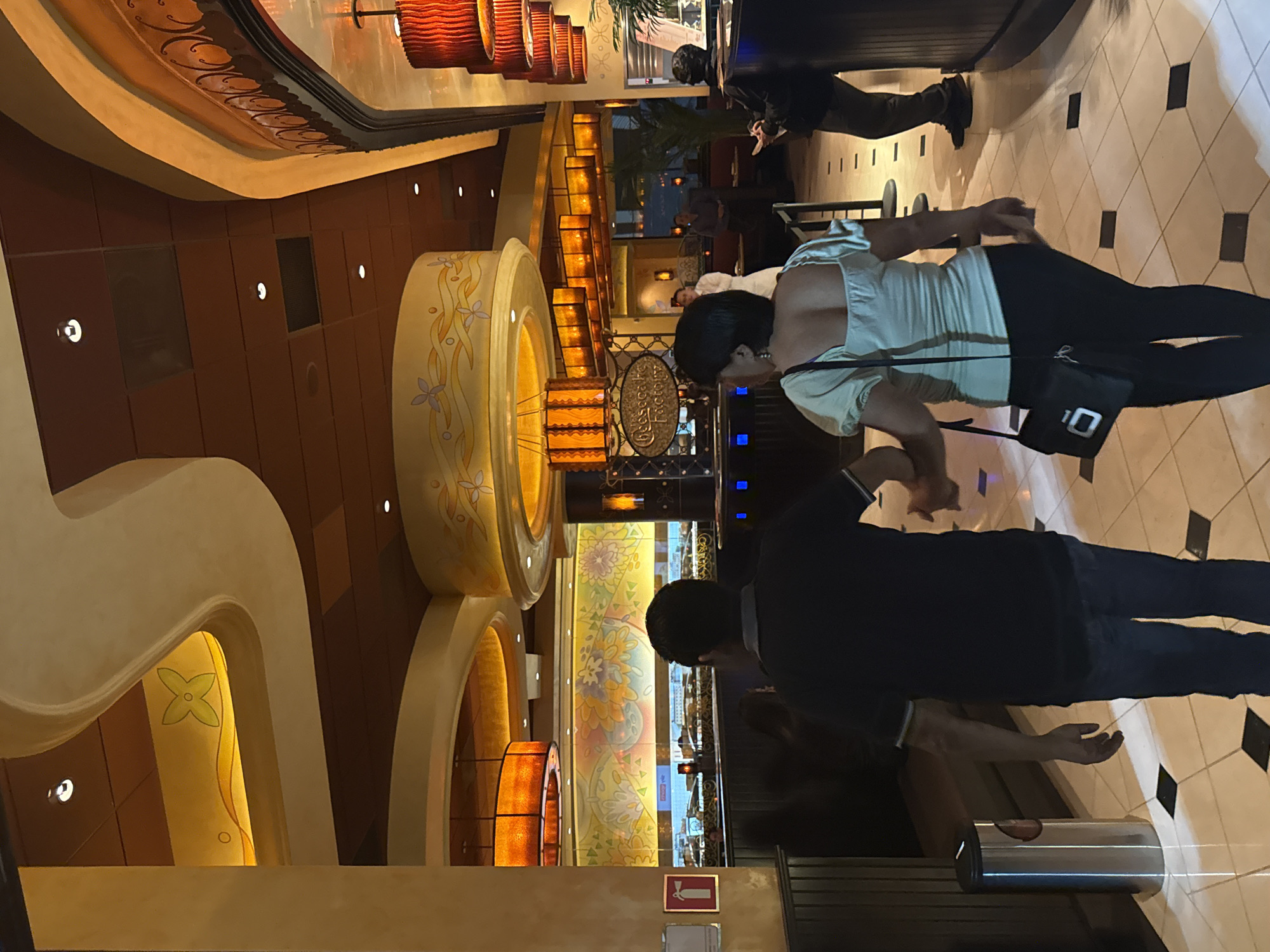 The interior of The Cheesecake Factory restaurant in Puebla, Mexico, is depicted in this vertically oriented image. Two individuals, a male and a female, stand in the foreground. The male wears a dark shirt and jeans, while the female wears a light-colored top and dark pants, with a black bag on her shoulder. They appear to be looking at something in the restaurant.
Behind them, the restaurant's interior features decorative elements such as large, circular, floral-patterned light fixtures and arched architectural details. The restaurant sign is visible in the background. Other patrons are seen seated at tables in the dimly lit restaurant. The floor is tiled in a black-and-white checkerboard pattern.