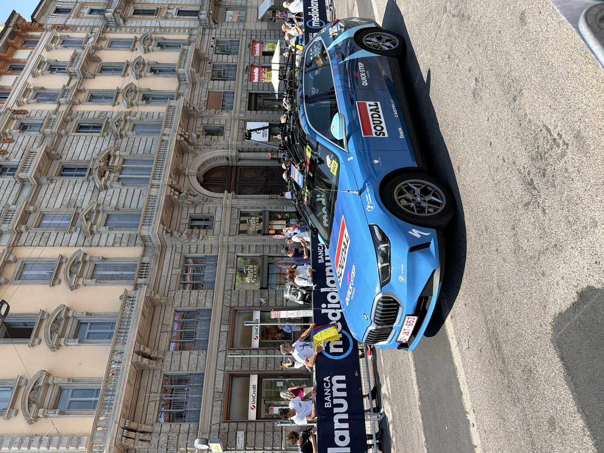 The image depicts a blue BMW car parked on a street in Milan, Italy. The car is adorned with logos including "Soudal," "Quick-Step Floors," and "Castelli." It has a license plate that reads "2-JAT-653." The car is positioned next to a "Banca Mediolanum" barrier. Several people are seen standing behind the barrier, appearing to be spectators.
In the background, there is an ornate multi-story building featuring intricate architectural details and several windows. Below it, there are storefronts with signs for businesses like "Unicredit" and "Nashi Salon." The street appears to be paved and well-lit, suggesting it is daytime. The overall setting is urban, with a mix of commercial and residential elements visible.