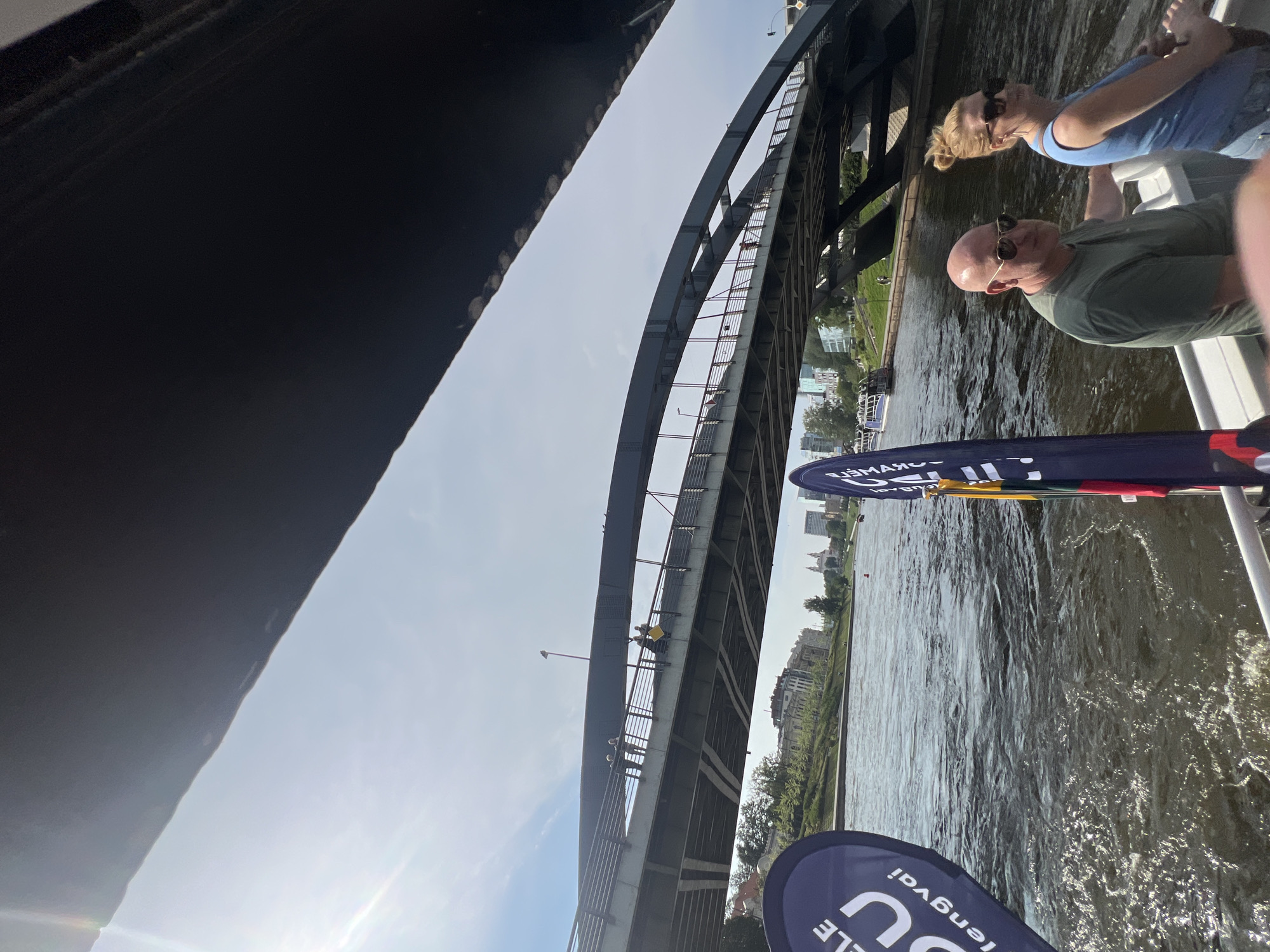 This is a ground-level, eye-level, wide shot of a body of water, likely a river in Vilnius, Lithuania, with a modern arch bridge spanning across it. The foreground is dominated by the rippling surface of the water, reflecting the bright, sunny sky. Two people are seated on a boat in the lower right quadrant of the image. The man, wearing a green t-shirt and sunglasses, is looking towards the camera. The woman, in a blue tank top and sunglasses, is looking to her right. Two dark blue banners with white text are visible on the left and right sides of the boat. The bridge is a prominent feature in the midground, with several people walking across it and architectural structures visible along its span. In the background, the cityscape of Vilnius can be seen, with buildings and trees lining the riverbank. The overall impression is a bright, daytime scene of urban life and leisure on the water.