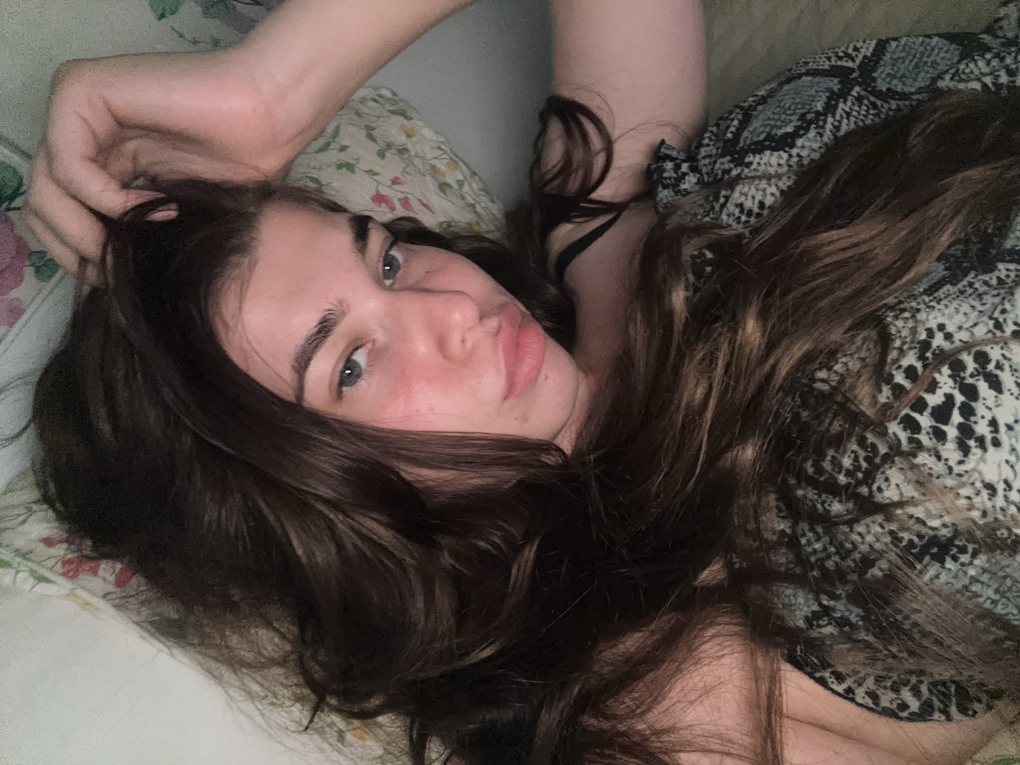A young woman with long, dark brown hair lies on a bed, propped up against a floral-patterned pillow. Her hand is resting on her head, fingers tangled in her hair. She is wearing a black top with a snakeskin-like pattern. Her face is turned towards the camera, with her eyes open and looking forward. Her cheeks are flushed, and her lips are slightly parted. The background is a soft, diffused light.